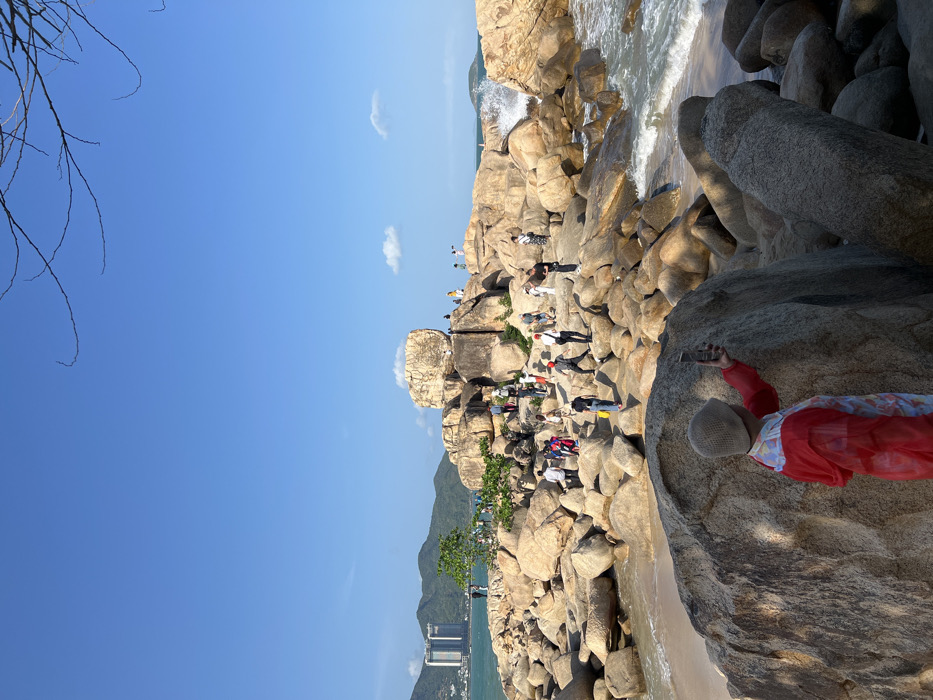 The image displays a sunny coastal scene at Hon Chong Promontory in Nha Trang, Vietnam. The foreground features a person from behind, wearing a light-colored beanie and a translucent red hooded jacket over a patterned top, holding a black smartphone vertically to capture the view. This individual stands on a large, textured grey boulder.
The central and midground are characterized by an extensive arrangement of numerous light-brown and grey granite boulders of varying sizes, forming a rocky promontory. Approximately 30 to 40 individuals are observed scattered across these rocks, engaged in walking, standing, and observing the surroundings. Some are positioned on higher boulders, while others navigate lower sections. Patches of light-colored sand are visible between some rock formations, transitioning into the ocean on the right. Waves are actively crashing against the rocks on the right side, producing visible white sea spray.
In the background, distant green hills and mountains rise from the left side of the coastline. Several modern multi-story high-rise buildings are discernible against the mountainous backdrop. The sky is clear and bright blue, with two small, scattered white clouds present. Sparse, bare tree branches extend into the upper right corner of the frame.