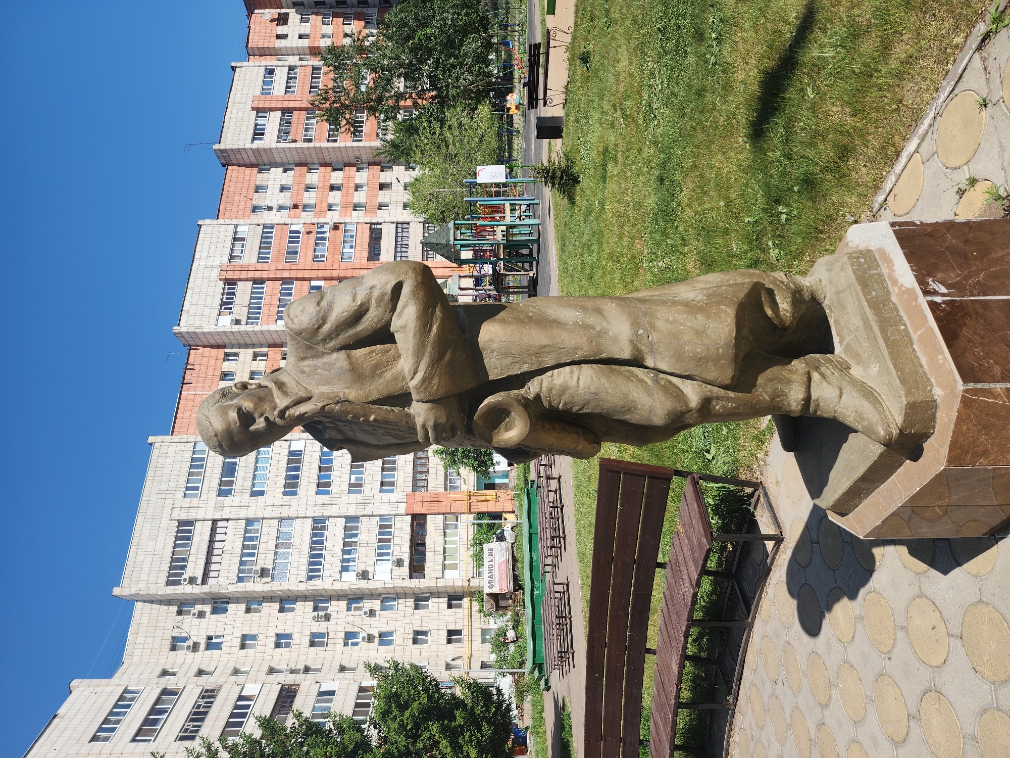 The image depicts a full-length statue of a man, likely a historical or cultural figure, situated in an outdoor public space in Kazan, Russia. The statue is sculpted in a light brown or gray material, likely stone or concrete, and stands on a multi-tiered base. The man is wearing a long coat and holds what appears to be a scroll or document.
In the background, there is a tall, multi-story apartment building with a light-colored facade, red accents, and numerous windows. There are trees and green lawn areas visible around the statue, as well as benches for seating. In the distance, playground equipment can be seen. The statue is on a paved area with a circular pattern. The sky is a clear blue.