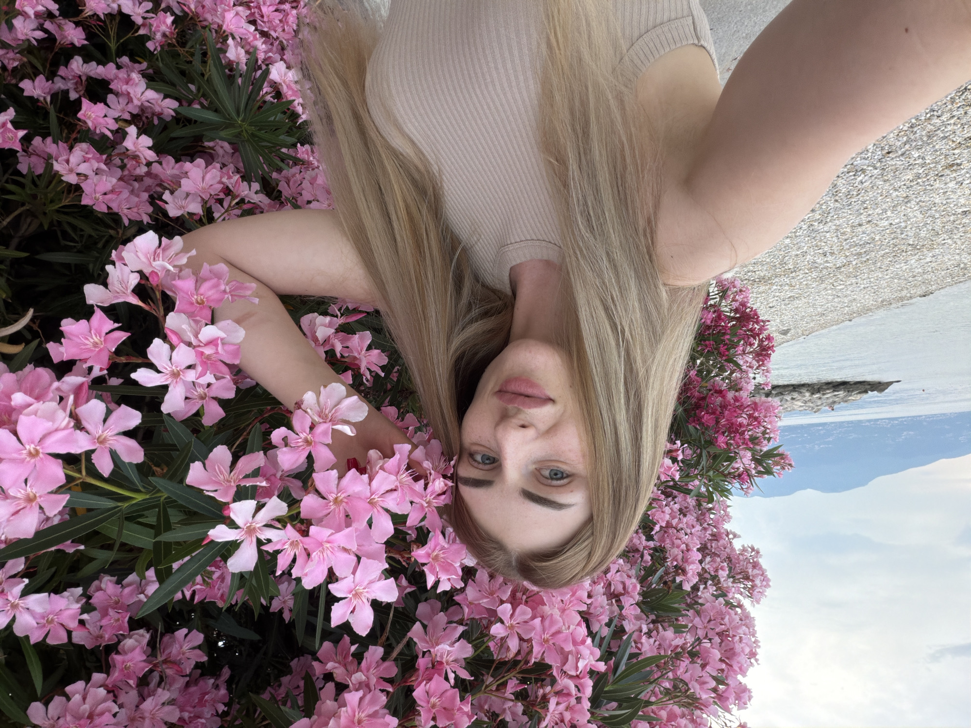 An outdoor, chest-up selfie shows a Caucasian woman with long, straight blonde hair and blue eyes. She is positioned to the left of the frame, with her body angled towards the right. She is wearing a light beige, ribbed tank top. Her right arm is extended, and her hand rests on a large cluster of pink oleander flowers that fill the right and upper portions of the image. Her left arm is out of frame.
Behind her, to the left, is a view of the sea, a rocky shoreline, and distant, hazy mountains under a cloudy sky. The ground in the foreground, to her left, is covered in small white and grey pebbles, suggesting a beach or waterfront. The overall scene conveys a summery, coastal atmosphere.