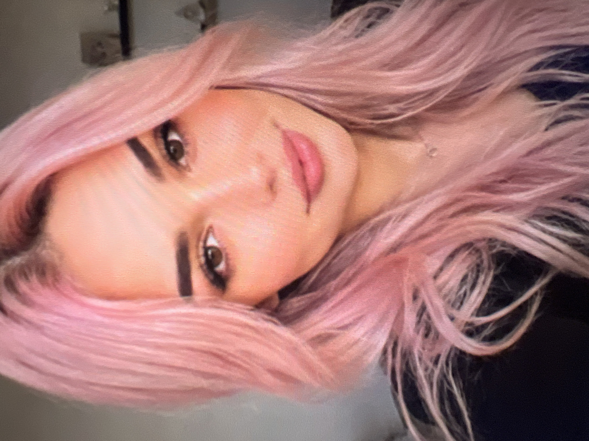 A headshot captures a young woman with striking pink hair. Her hair, dyed a vibrant pastel shade, falls in soft waves around her face, framing her features. She has defined eyebrows that arch gracefully over her expressive brown eyes. Her lips are a soft pink, appearing to be lightly glossed. She is wearing a dark top, and a delicate necklace can be seen resting on her collarbone. The background is blurred, suggesting an indoor setting, possibly a room with shelves and decorative items visible in the distance. The lighting appears to be soft and natural, highlighting the contours of her face.