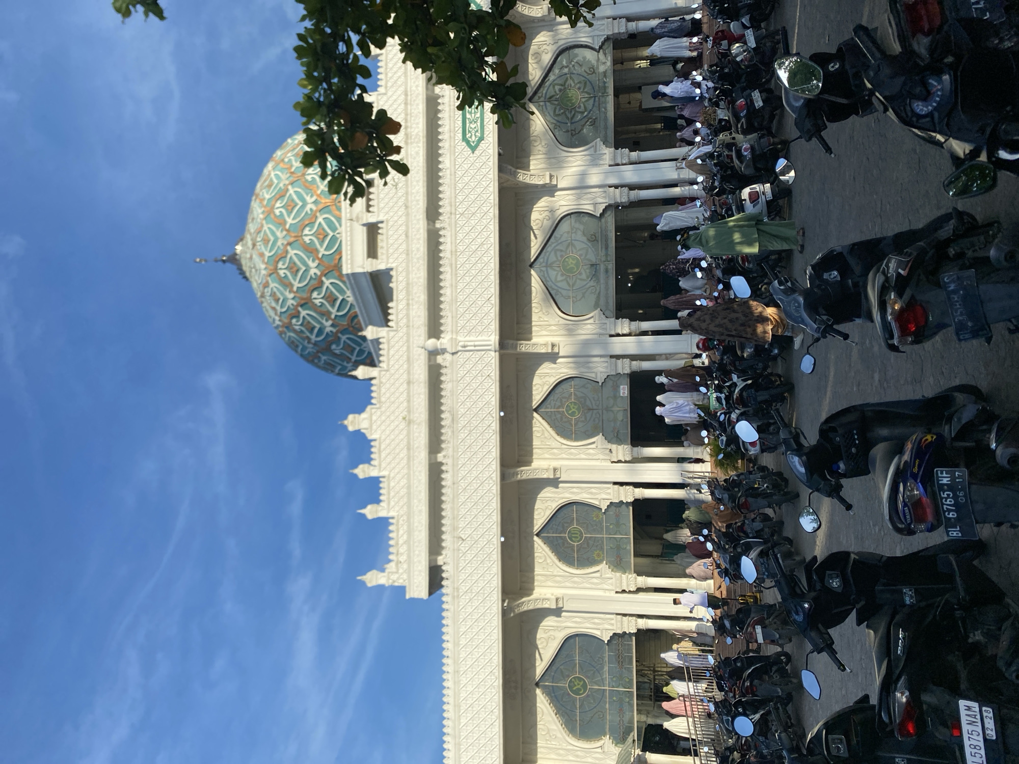 The image shows a mosque in Lhokseumawe, Indonesia, during prayers. The mosque has a blue and gold dome, white walls, and arched windows. People are praying inside the mosque and on the porch in front of the mosque. Many motorcycles are parked in front of the mosque. The sky is blue with some clouds, and there are tree branches visible at the top of the frame.