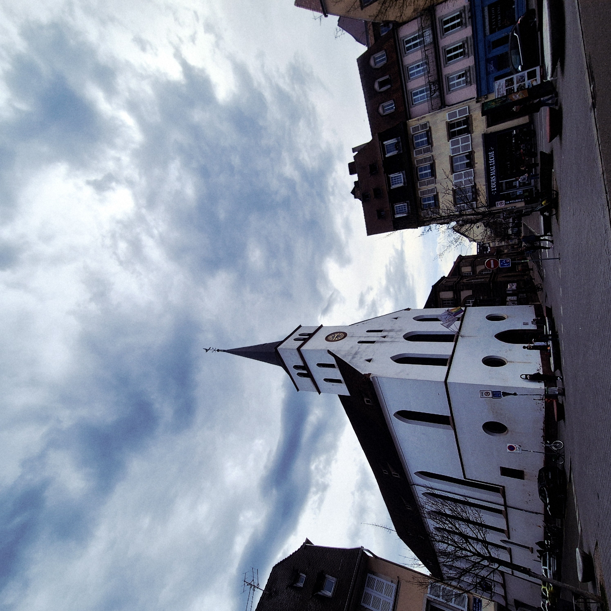 The image presents a low-angle view of a street in Strasbourg, France, focusing on the Église Saint-Pierre-le-Jeune. The white church, with its spire and visible clock, is juxtaposed against a dramatic sky filled with wispy clouds. Adjacent to the church is a street lined with traditional European buildings, some displaying colorful facades, including pink, yellow, and blue hues. A few pedestrians are discernible on the right side of the street, next to a row of parked cars and bicycles. The street is paved with cobblestones. Bare trees are visible along the street, indicating it could be late fall or winter. Signposts and signage are present.