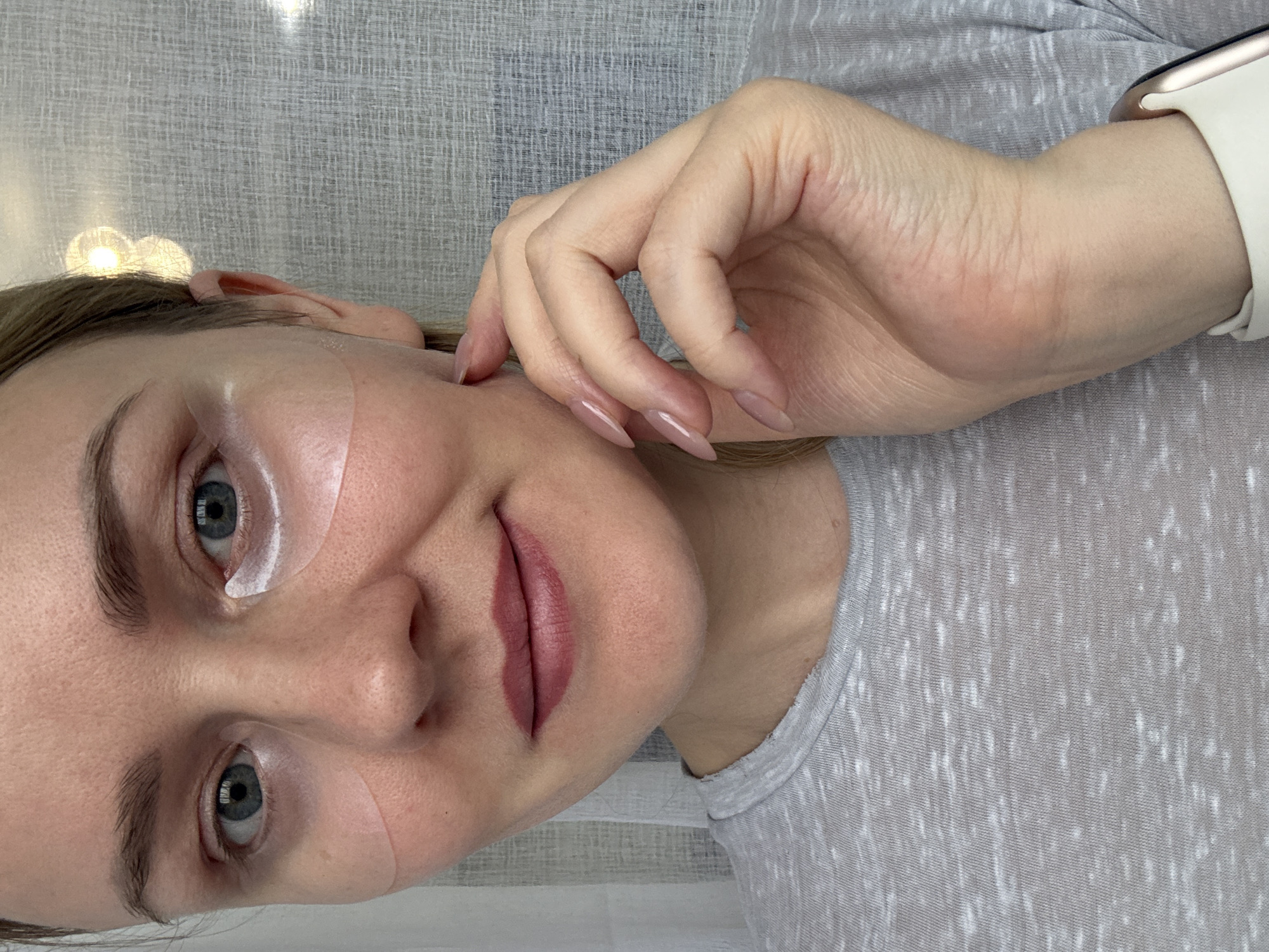 The original image is rotated 90 degrees clockwise, showing a close-up of a woman's face and upper body, appearing to be in Chaikovsky, Russia. The woman, with light skin, blue eyes, and light brown hair, is looking directly at the camera with a neutral to subtly content expression.
She appears to be undergoing a beauty treatment, as evidenced by two white, translucent under-eye patches placed beneath her lower eyelids; one is fully visible under her left eye (from her perspective), and the other partially under her right eye. Her lips are colored with a muted, reddish-pink tint. Her left hand rests gently near her face, showcasing long, well-maintained fingernails painted with a light nude polish. She is wearing a light grey, mottled short-sleeved or long-sleeved top with a crew neck. On her left wrist, a smartwatch with a white band, resembling an Apple Watch, is visible.
The background consists of a textured light grey or white surface, possibly wallpaper or a curtain. In the upper left corner of the original image (which would be above her head if oriented correctly), there are a few soft, out-of-focus light sources. The scene is indoors, and the time of day is not discernible. No visible text is present in the image.
