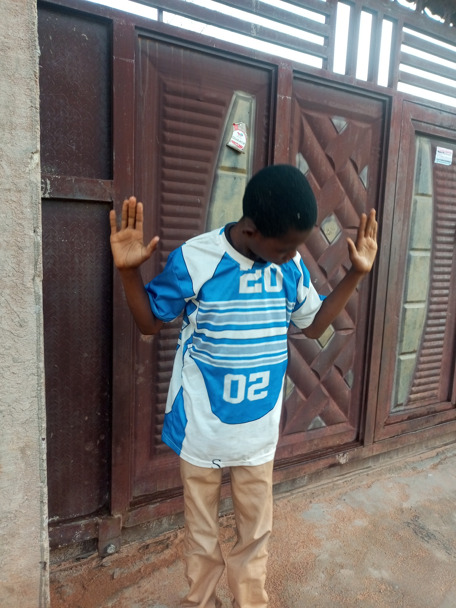 A young Black male wearing a white and blue horizontally striped t-shirt with the numbers "20" and "02" printed on it, and tan pants, stands with his hands raised in front of a dark brown double gate with a diamond plate insert on the left side. The gate is located on the side of a building in Tempure, Nigeria. The gate has decorative V-shaped ridges. A white sticker with "QUARTZ" and a red and white tag are attached to the left side of the gate. The ground in front of the gate is sandy and reddish-brown. The building wall to the left of the gate is textured and light brown. The sky is visible above the gate through a metal grate.