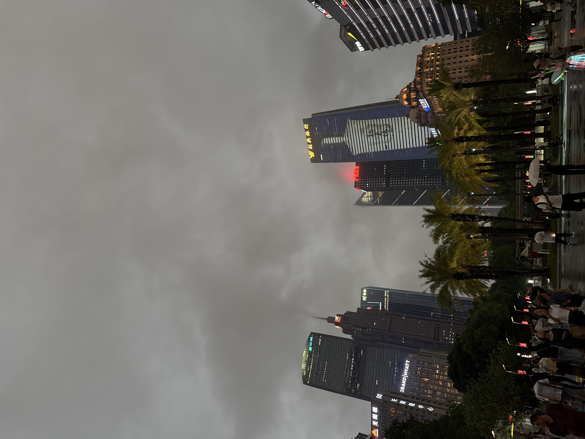 The image shows a street scene in the Tianhe District, China, on an overcast day. Skyscrapers are visible, illuminated with neon signs and lights, piercing through the heavy cloud cover. Palm trees line a street bordering the skyscrapers, and pedestrians are present on the sidewalk.