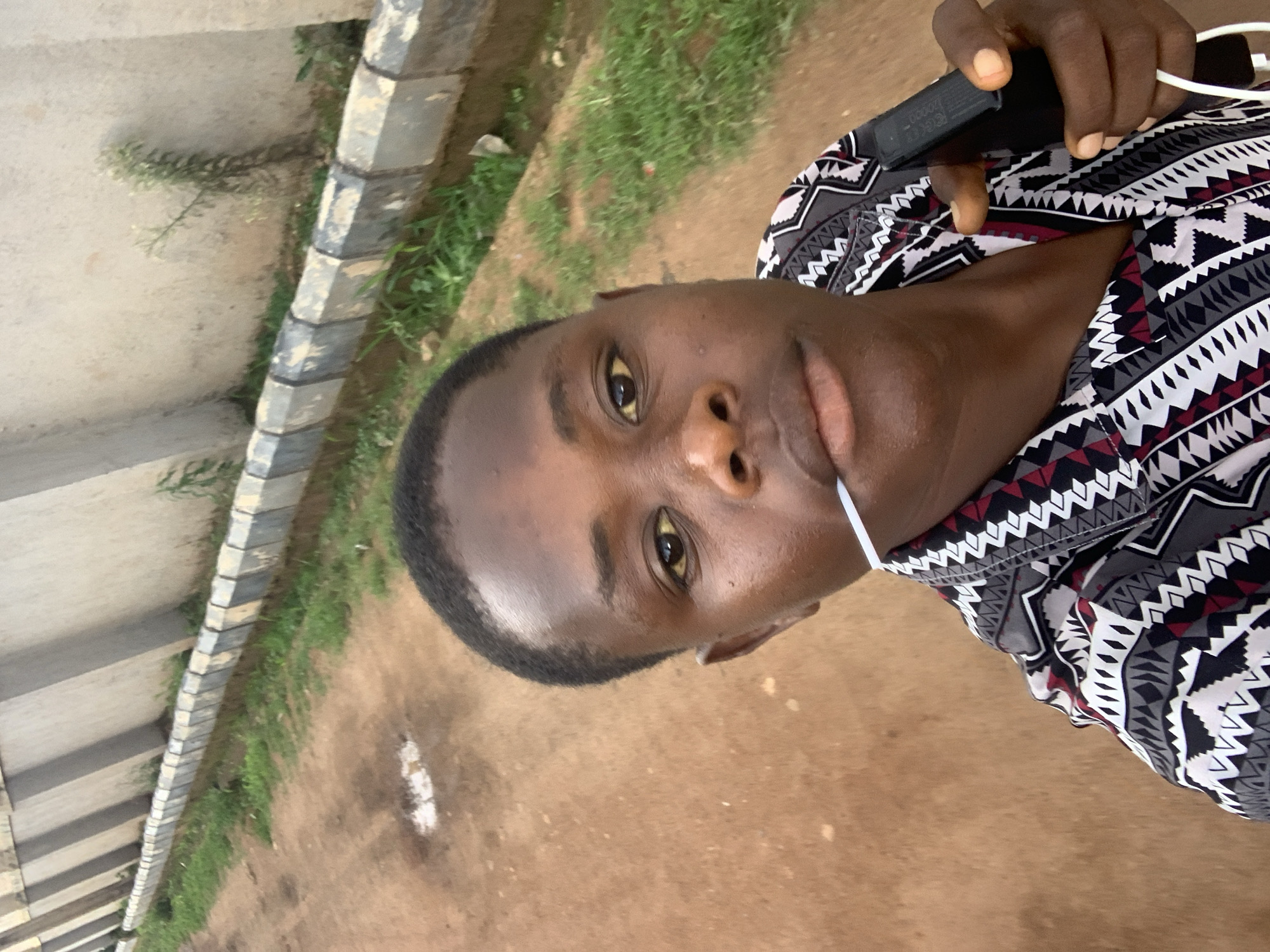 This horizontally oriented image features a close-up portrait of a young person, likely male, with dark skin and short dark hair, looking directly at the camera. Their expression is neutral, and a white string (possibly from a face mask or headphones) is visible near their mouth.
The person is wearing a shirt adorned with a distinct geometric pattern in shades of black, white, gray, and maroon. In the upper right quadrant of the image, their hand is seen holding a black rectangular device, possibly a smartphone or portable player, from which white wires (likely earbuds or headphones) extend. The text "CG C20000-1" is legible on the black device.
The background suggests an outdoor location in Gura Top, Nigeria. To the left, a concrete structure, possibly part of an overpass or barrier, is visible, featuring alternating light and dark gray sections. Green foliage and patches of grass grow along the base of this structure and on the reddish-brown, sandy ground that dominates the rest of the background. The scene is illuminated by natural daylight.