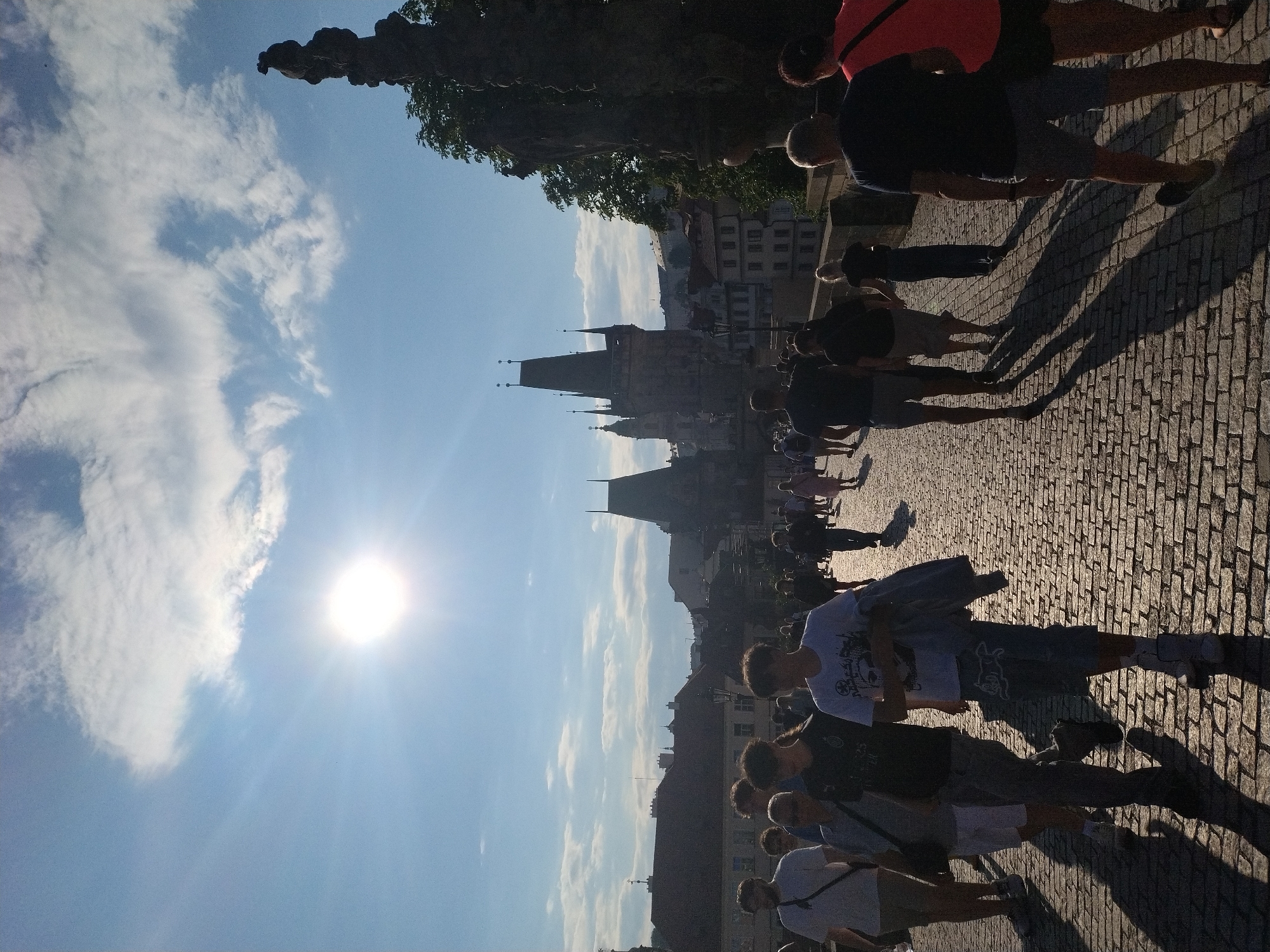 The image shows a crowded cobblestone bridge in Prague, Czechia, under a bright, sunny sky with scattered clouds. The sun is high in the sky, casting long shadows. People of various ages are walking in both directions on the bridge, which is lined with statues on the right side. In the background, historical buildings and towers are visible, including a distinctive gothic tower with spires. The overall impression is a vibrant, busy scene on a historical landmark in Prague.