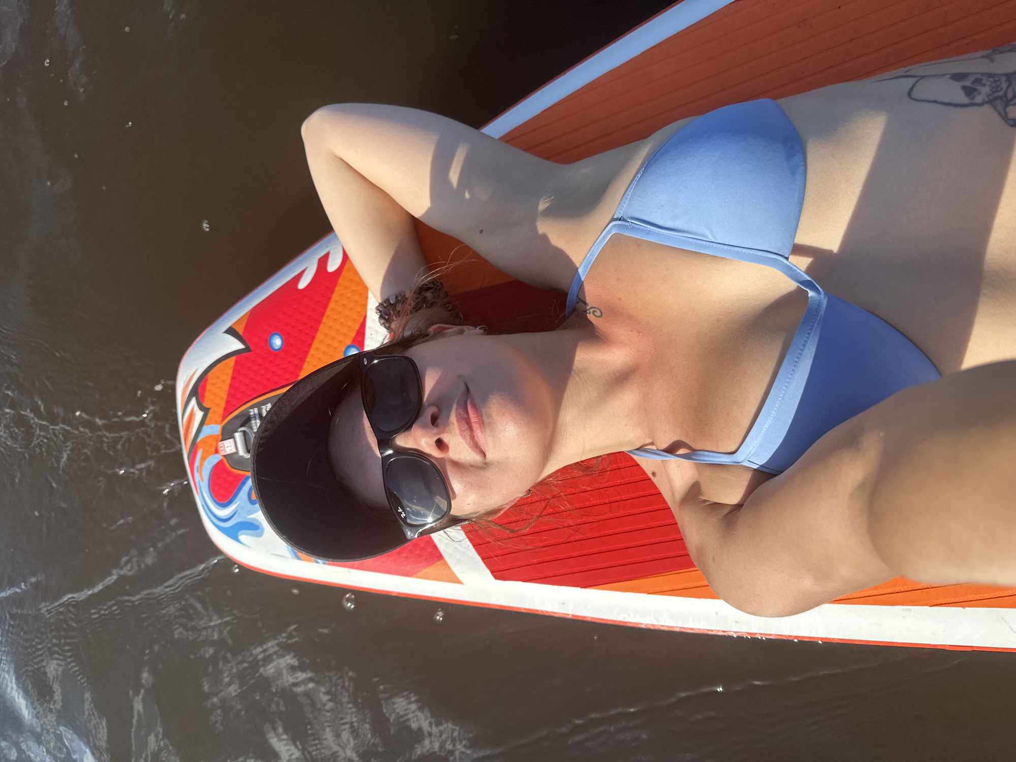 A woman is lying on a paddleboard in dark water. She is wearing a blue bikini top, sunglasses, and a black baseball cap. Her right arm is behind her head. The paddleboard is orange, white, and red with a graphic design on the front. The water is choppy and appears to be reflecting sunlight. There is a tattoo of a skull on her left arm. The scene is likely outdoors, possibly on a lake or river in Mikhalevo, Russia, given the user's request.