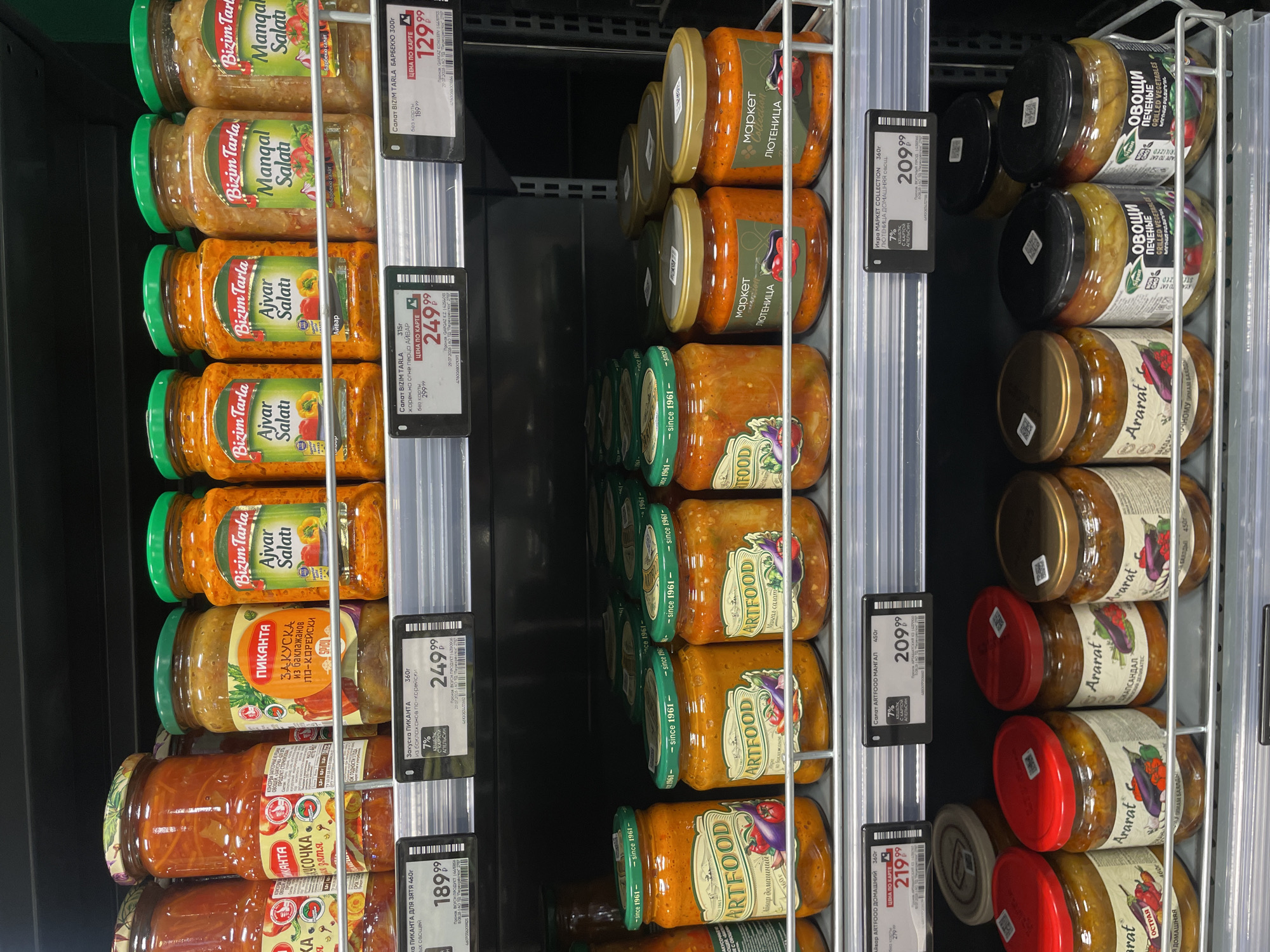 The image shows a supermarket aisle in Pargolovo, Russia, specifically within a store likely belonging to the "Lenta" hypermarket chain, indicated by the price tags and shelf design. The focus is on a display of preserved vegetables in jars, primarily on two shelves. The top shelf features several brands, including "Bizim Tarla" with "Ajvar Salati" and "Manqal Salati" varieties, as well as "Picanta" with a vegetable spread. Jars of "Ararat" brand vegetable preserves are visible on the bottom shelf, alongside "Artfood" and "Market Collection" products. The lighting is artificial, typical of an indoor retail environment, and the overall scene is clean and organized. There are no people visible in the image.