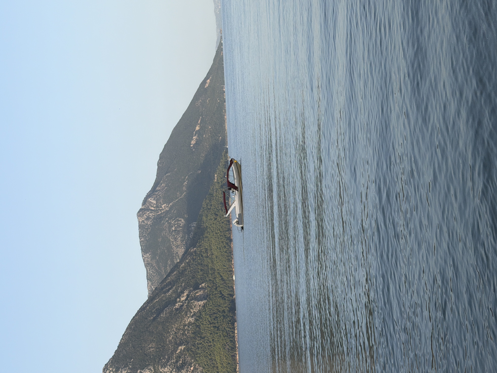 A motorboat with a red canopy floats on calm, blue water. Behind the boat, a tree-covered mountain range slopes down to the water's edge. The sky above is clear and blue. The scene is located in the Municipal Unit of Alyzia, Greece.