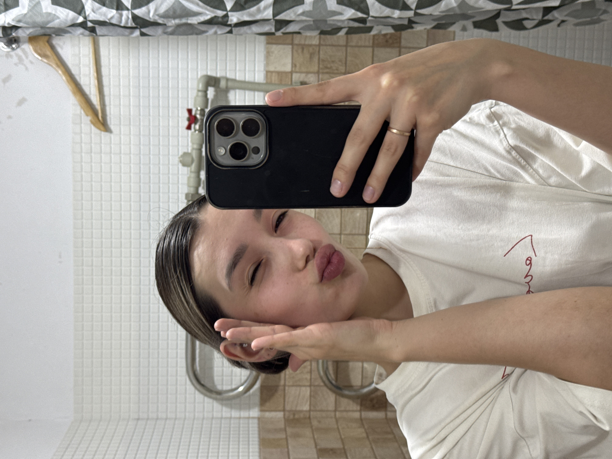 A person is captured in a mirror selfie, holding a black smartphone with a triple camera array. The individual, appearing to be female, is shown from the chest up, making a playful expression with puckered lips and one eye winking shut. Her left hand is posed under her chin, while her right hand holds the phone. She is wearing a white t-shirt, which has a partially visible red design or text on the left chest area, starting with a stylized upward stroke followed by a curly line. A simple gold-colored band ring is visible on the ring finger of her right hand.
The background, a reflection in the mirror, shows elements of a bathroom. A white shower curtain with a black and white geometric pattern hangs at the top. The walls are covered in square tiles: white tiles on the upper left and a section of brown/beige tiles on the lower right. Plumbing fixtures, including silver pipes and a red valve, are visible on the right side of the reflection. A wooden clothes hanger hangs from a rail on the left, and curved chrome-like metal bars are present in the lower background, possibly towel rails or shower grab bars. The lighting appears to be indoor, and the time of day is not discernible. The overall mood conveyed is lighthearted and casual.