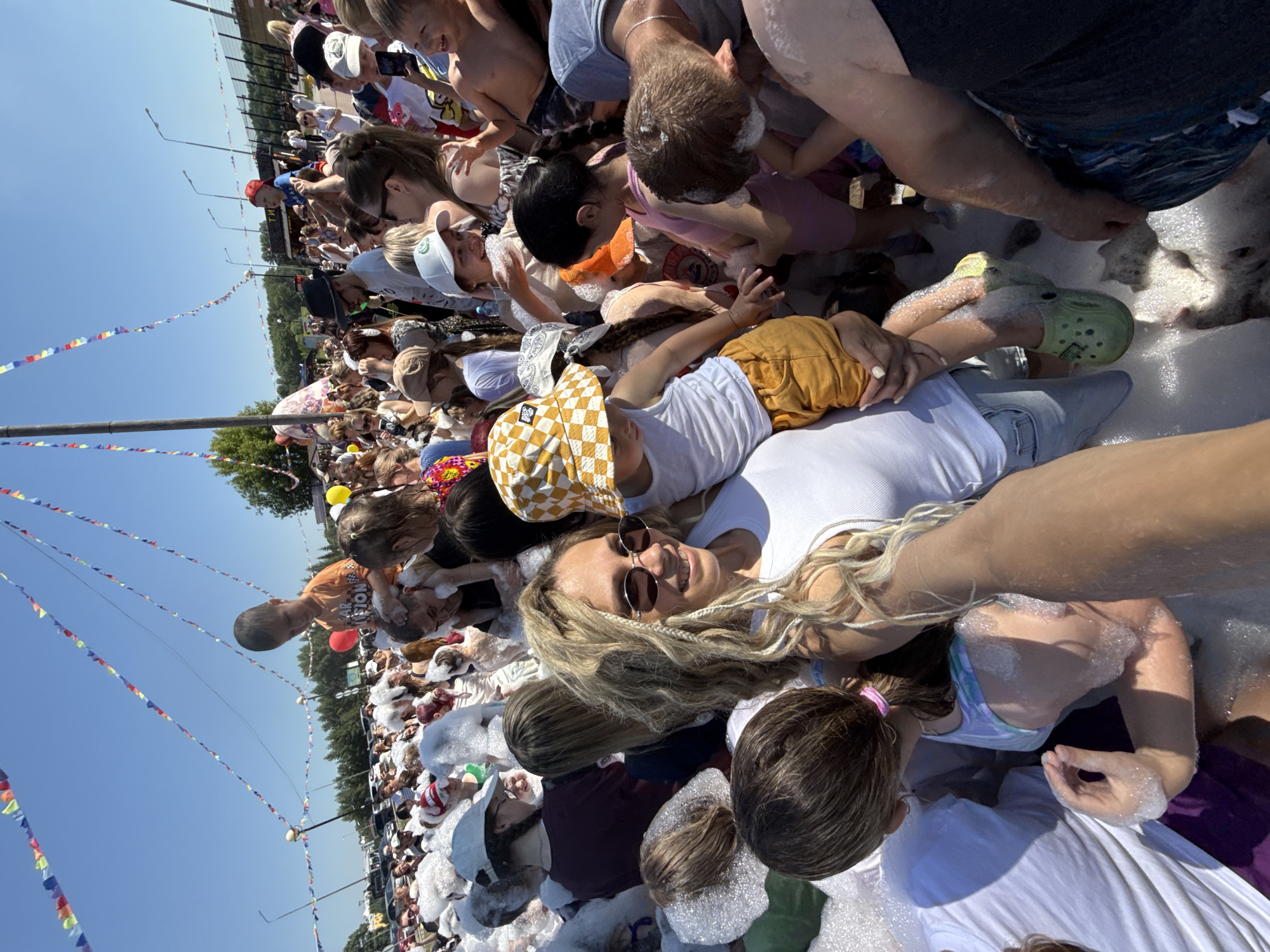The image shows a large outdoor gathering of people, many of whom are children, participating in a foam party on a sunny day. The setting appears to be an open area, possibly a park or festival ground, with trees and buildings visible in the background. Colorful triangular flags are strung across the sky, suggesting a festive event. The ground is covered in a thick layer of white foam, with many individuals immersed in it. People are interacting, some are being held up by others, and children are playing in the foam. A woman in the foreground is taking a selfie with a child sitting on her shoulders. The atmosphere is lively and joyful. The scene is located in the city of Zavolzhye, Russia.
