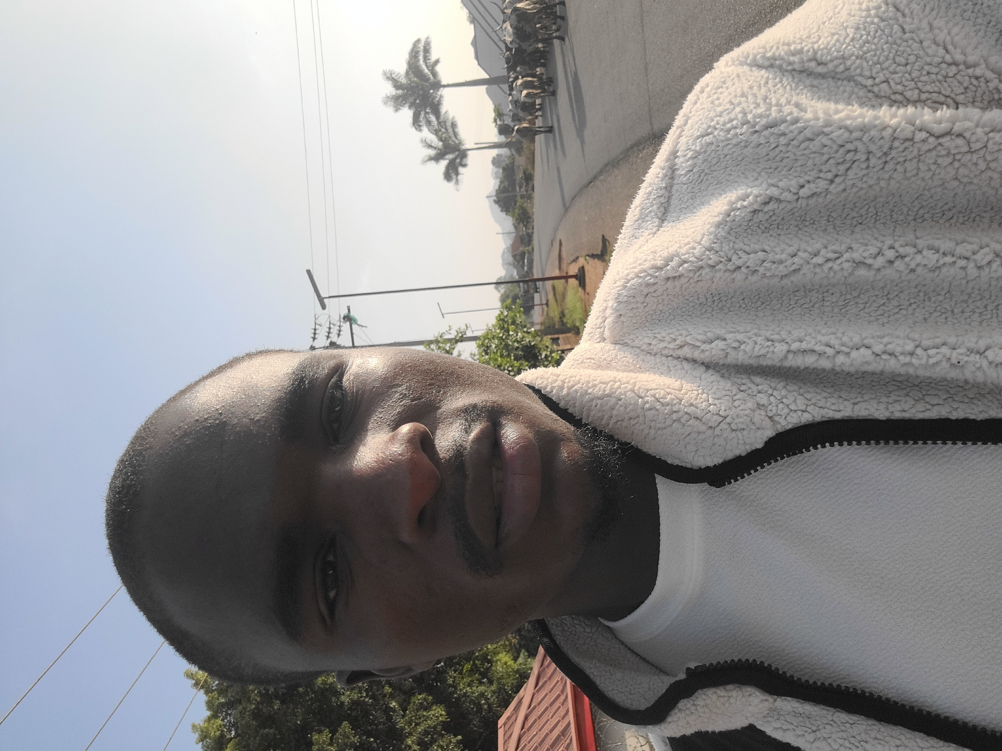 A vertical low-angle shot depicts a man in the foreground and an outdoor scene in the background. The man has dark skin, a closely shaved head, and a small beard. He wears a white t-shirt and a white fleece jacket with a black zipper.
The background consists of a street with palm trees and a group of animals, possibly livestock, crossing the road. There are utility poles with wires visible, as well as greenery and a building with a red roof. The sky is light blue with some haze.