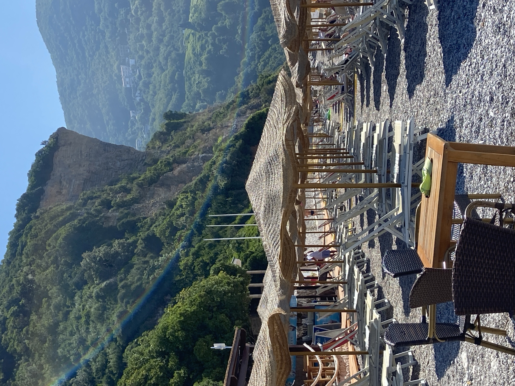 The image depicts a sun-drenched beach in Prijevor, Montenegro, with a dramatic cliff face rising behind it. Rows of empty lounge chairs and thatched umbrellas stretch across the pebbled beach, suggesting a leisurely atmosphere. A rainbow arcs across the lush green hillside, adding a touch of natural wonder to the scene. In the foreground, a wooden table with two chairs stands ready for patrons, with a single green object resting on its surface. A few individuals are visible in the middle ground, enjoying the beach, while the background reveals a hazy view of buildings nestled amongst the verdant hills. The clear blue sky and bright sunlight indicate a warm and pleasant day.