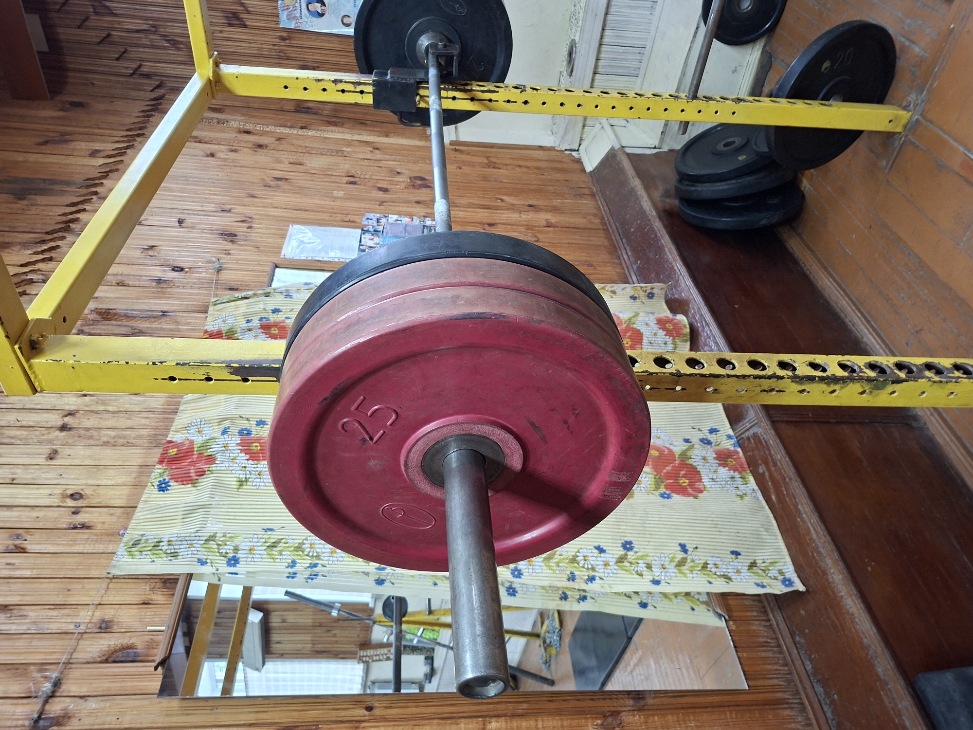 The image shows a close-up view of a yellow weightlifting rack with a barbell loaded with red and black weight plates. The barbell is positioned horizontally across the rack. One of the red weight plates is clearly marked with "25". In the background, a wooden paneled wall is visible, adorned with a patterned curtain and a mirror. A few black weight plates are stacked on the floor to the right of the rack. The overall environment appears to be an indoor gym. There is no discernible indication of the specific location in Kazan, Russia, within the image itself.
