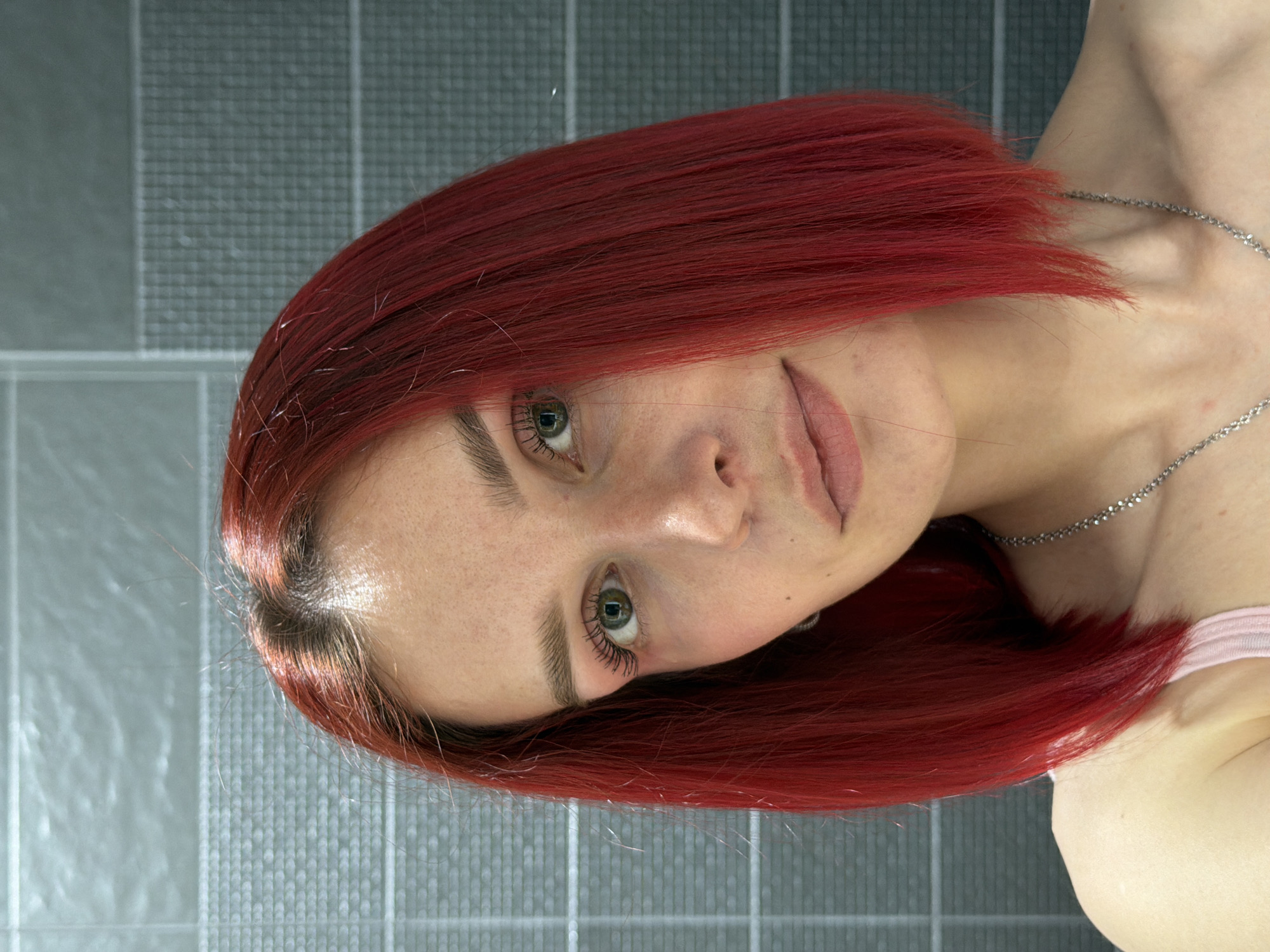 A head-and-shoulders portrait shows a fair-skinned person with red, shoulder-length hair, green eyes, and a silver necklace against a grey tiled background. The person is looking towards the camera and angled slightly downward. The lighting is even. There are no discernible details suggesting the location is Chaikovsky, Russia.