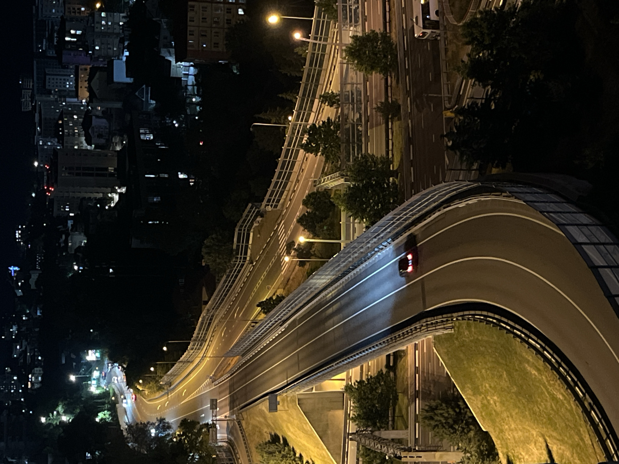 This is an aerial night photograph of a multi-level highway interchange in Sochi, Russia. The interchange is illuminated by streetlights and the headlights of vehicles. In the foreground, a red car drives on an elevated curved section of the highway, its brake lights illuminated. Below this, another section of the highway is visible with multiple lanes and cars. In the background, the city of Sochi is visible with numerous buildings and lights. The scene is framed by dark trees and the night sky.