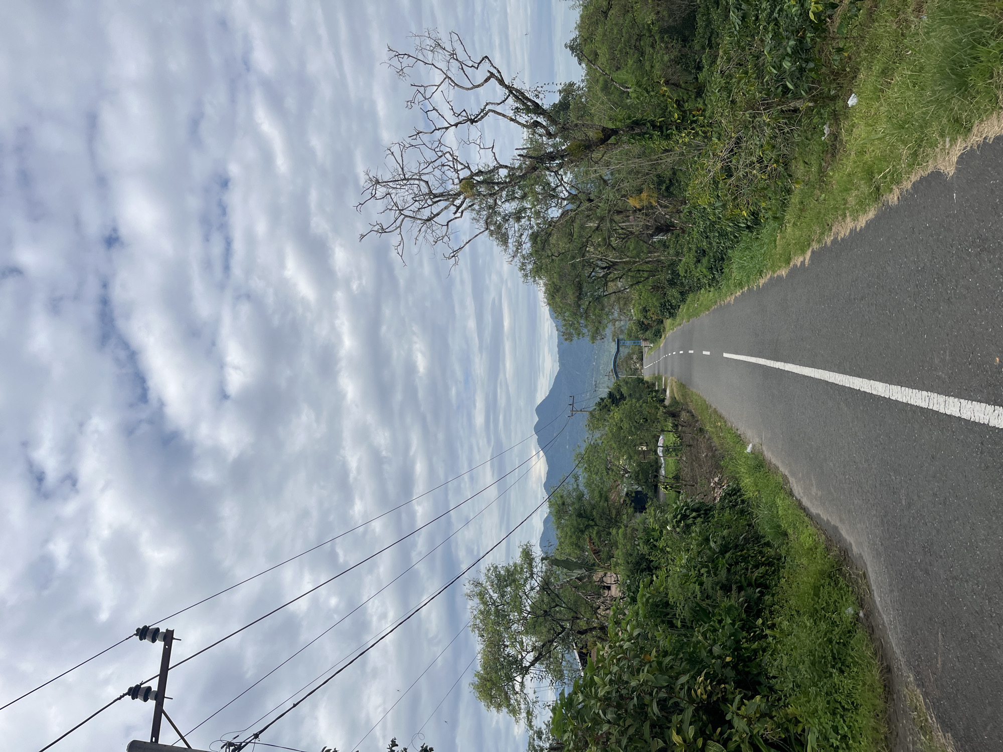 A straight, paved road with a white dashed line in the center stretches into the distance. The road is flanked by lush green vegetation and trees. In the background, mountains are visible under a cloudy sky. Electrical wires and poles are present on the left side of the image. The scene appears to be a rural area, possibly in or near Damaranbaru, Indonesia, given the presence of tropical foliage and mountainous terrain. There are no people or vehicles visible in the image.