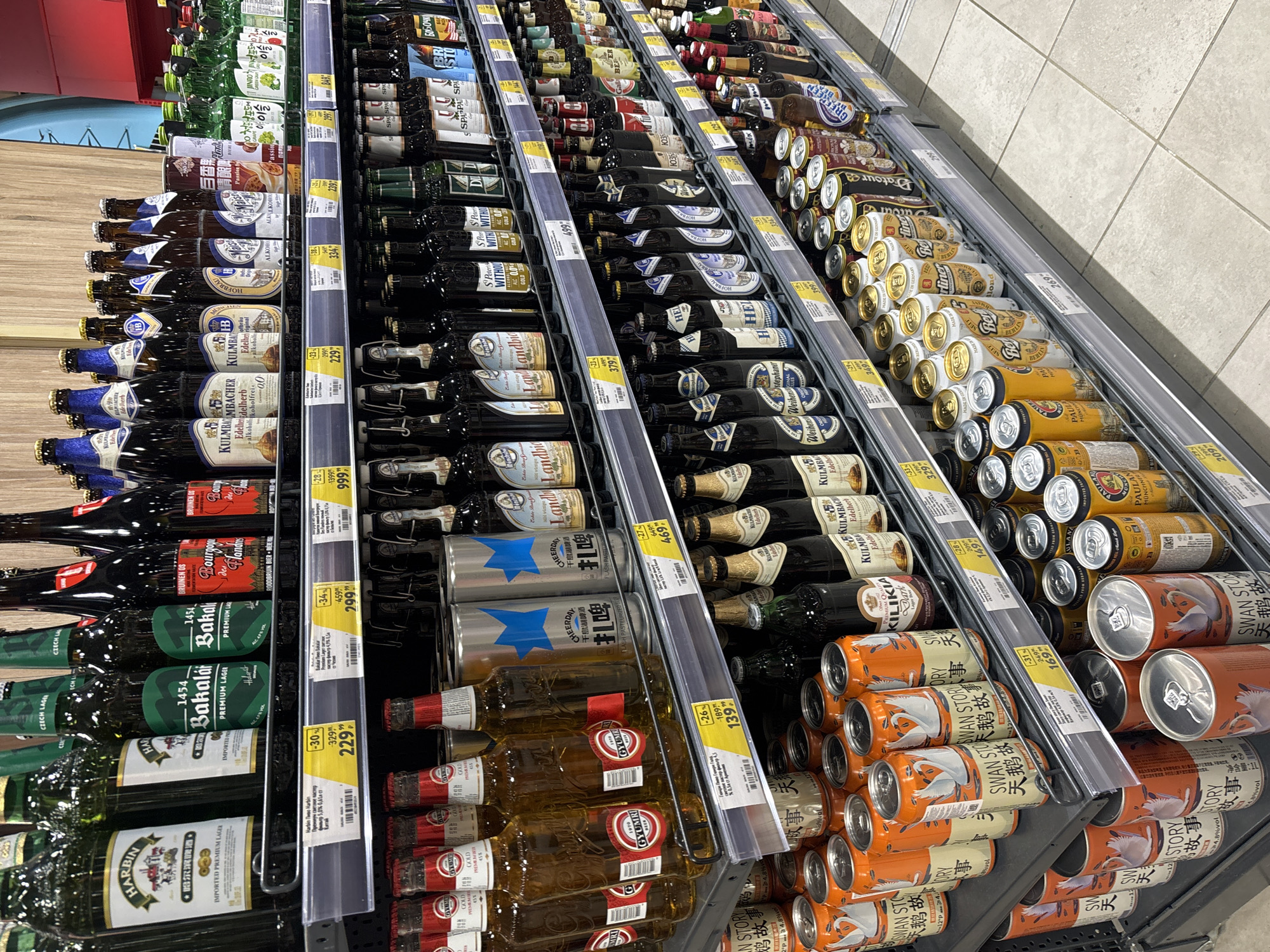 The image shows a refrigerated shelf in a store, stocked with a variety of bottled and canned alcoholic beverages, predominantly beer. The brands include local brews alongside international labels, many in Chinese and German languages. Prices in Russian rubles are displayed on shelf labels. The location is likely in a store somewhere in Russia, most plausibly the city of Sochi based on the product availability and price tags.