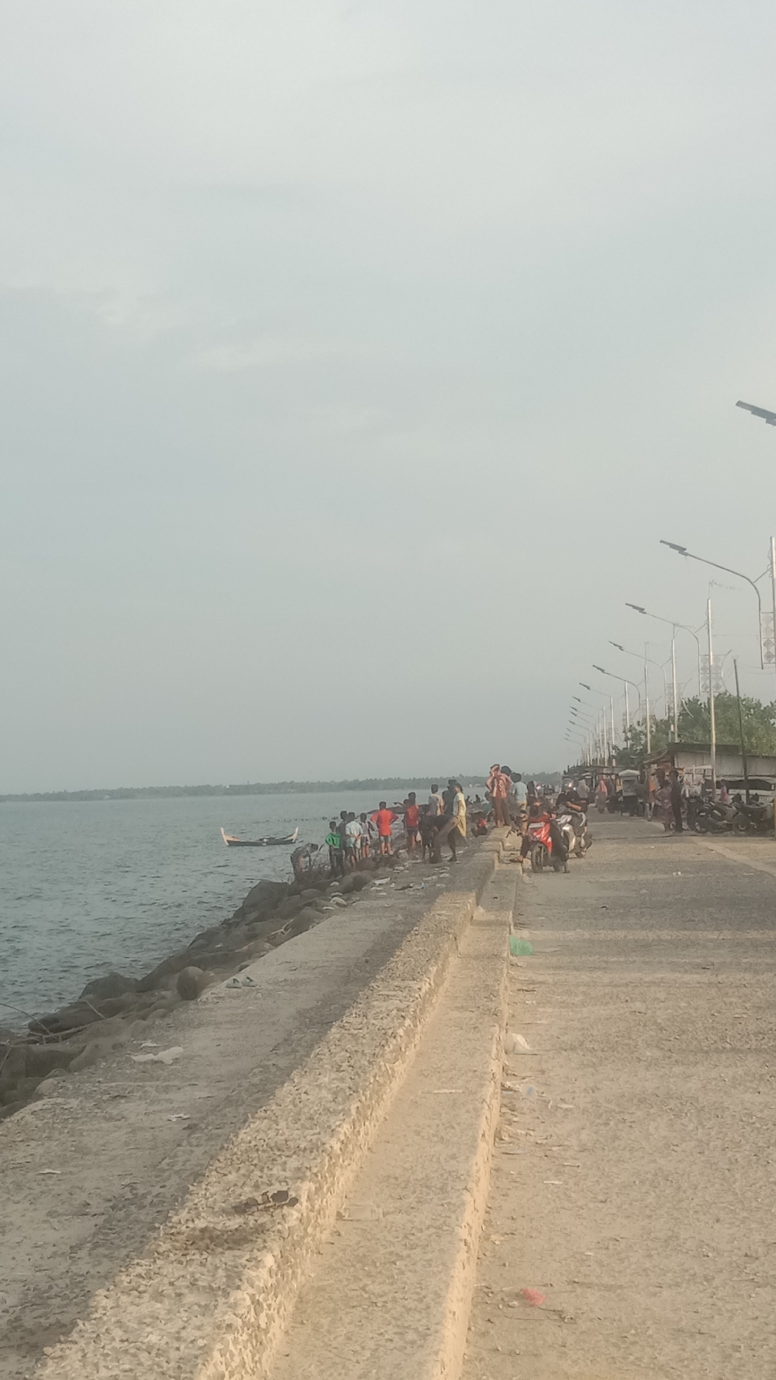 This image depicts a lively coastal scene, likely in Lhokseumawe, Indonesia, characterized by a busy waterfront area with numerous people, vehicles, and a calm body of water.
The foreground features a textured concrete barrier or seawall running horizontally across the image, separating a paved road from the water's edge. Along the water, there's a rocky embankment.
**Subjects and Activity:**
Many people are gathered along the concrete barrier and the rocky shore on the left side of the image, appearing to interact with the water. Some individuals are standing, while others are closer to the water's surface. One small, traditional-style wooden boat is visible in the calm water near this group. On the paved road to the right, a larger crowd of people is present, and several motorcycles are parked or moving, suggesting active commerce or commuting.
**Objects and Setting:**
The paved road is lined with a series of modern streetlights on tall poles that extend into the distance. In the far background on the right, glimpses of buildings or structures are visible behind trees, indicating a developed area adjacent to the coast. The water is calm and extends to a distant landmass on the horizon under a hazy or overcast sky.
**Notable Details:**
The sky is a uniform, light gray, suggesting an overcast day or significant atmospheric haze, which diffuses the sunlight and creates a soft, even lighting across the scene. The time of day appears to be daylight, possibly morning or late afternoon given the muted light. The overall atmosphere is one of activity and community engagement along the waterfront. No visible text can be discerned in the image.