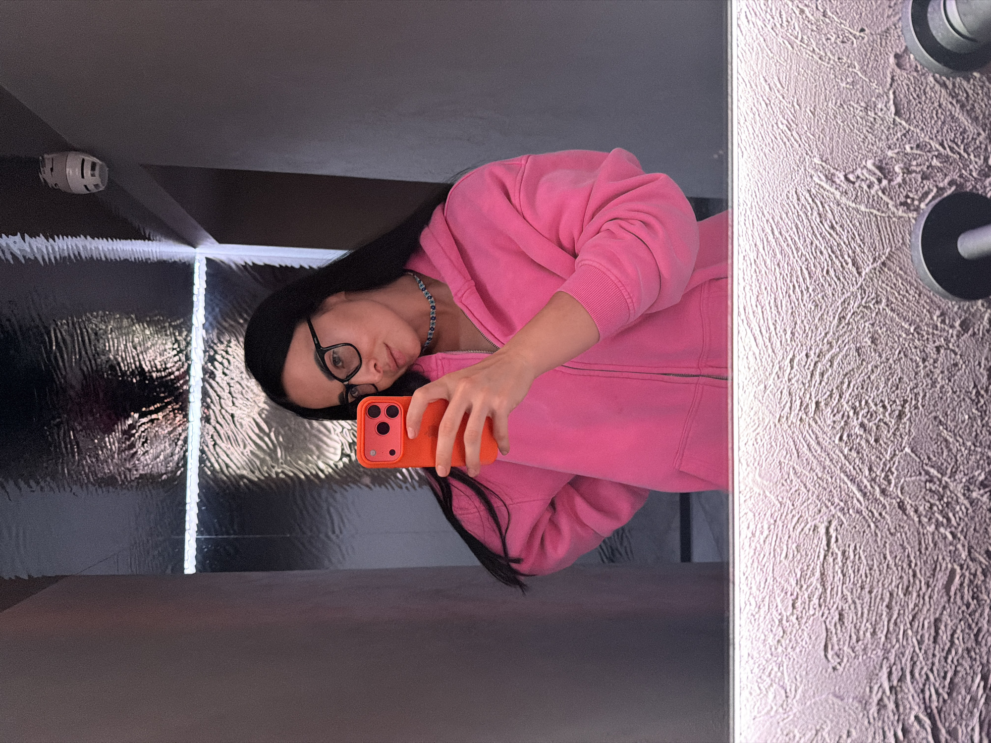 This image is a mirror selfie, rotated 90 degrees counter-clockwise, featuring a person in an indoor setting.
The main subject is a person with long, dark hair and black-framed glasses, wearing a bright pink hooded sweatshirt (hoodie) with a zipper and a light blue beaded necklace. They are holding an orange smartphone with a triple camera setup in their right hand, using it to take the picture. The person has a neutral expression, looking towards the camera.
The background on the left side of the frame shows a dark, highly reflective surface, possibly a large mirror or polished panel. This surface reflects a bright, vertical strip of light, likely an LED fixture, and a white, cylindrical device (possibly a smoke detector) mounted on a ceiling or upper wall. The reflection creates a sense of depth and modern design.
On the right side of the frame, a distinctively textured, light-colored wall is visible. This wall appears to be finished with a rough plaster or concrete texture. Two dark, cylindrical objects, resembling door handles or hooks, are mounted on this textured wall.
The scene depicts an interior space, characterized by modern lighting and varied wall textures, located in Saint Petersburg, Russia. The time of day is not discernible, as the setting is indoors. There is no visible text in the image.