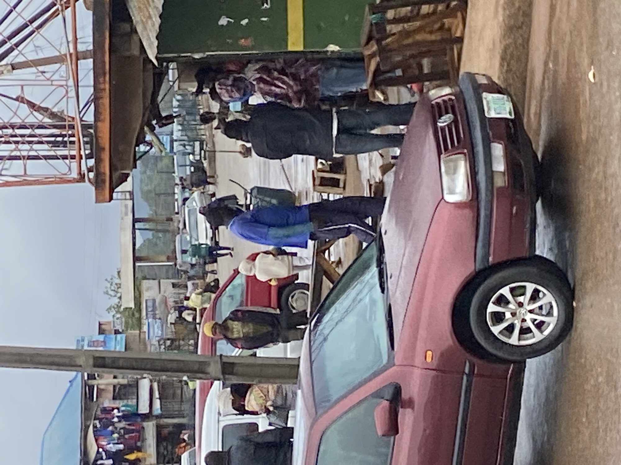 In Bukuru, Nigeria, on a cloudy day, a group of people are gathered near a street market. A maroon Volkswagen Golf car is parked in the foreground, facing left. Behind it, a white minivan and a red van are also visible. Several individuals are standing and walking around the vehicles, some carrying goods or engaging in conversation.
The market stalls display various items, including clothing and food. A tall metal structure, possibly a communication tower, looms in the background. The ground appears wet, suggesting recent rain. The overall atmosphere is one of everyday street life, with a sense of commerce and community.