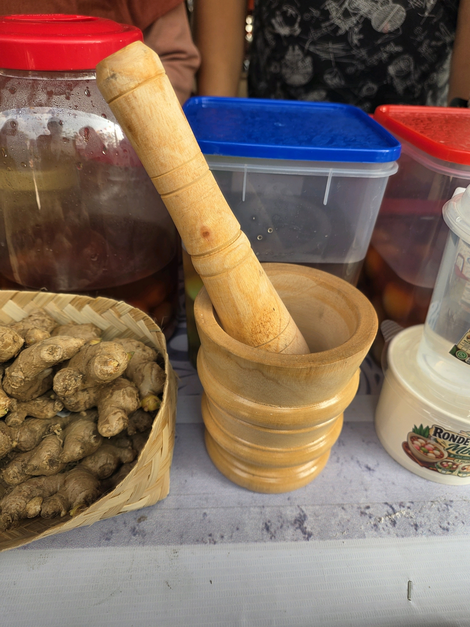 This is a close-up, eye-level, still life photograph taken outdoors on a table, likely in Bandung City, Indonesia. The scene is dominated by kitchen implements and ingredients. In the foreground, a woven bamboo basket filled with fresh ginger root is visible on the left. To the right of the basket, a wooden mortar and pestle are placed. The mortar is cylindrical with decorative rings along its body, and the wooden pestle is inserted into it. Behind the mortar and pestle, several large plastic containers with colorful lids are visible. The leftmost container has a red lid, the one next to it has a blue lid, and further to the right, another container with a red lid is partially visible. These containers appear to hold a dark liquid and possibly some fruit or other ingredients. A white tub with a label featuring illustrations of food items is positioned to the far right. The background is blurred, but suggests an outdoor setting with people partially visible, one wearing a patterned shirt. The overall impression is of a food preparation area, possibly for making traditional Indonesian drinks or dishes, given the presence of ginger and the style of containers.