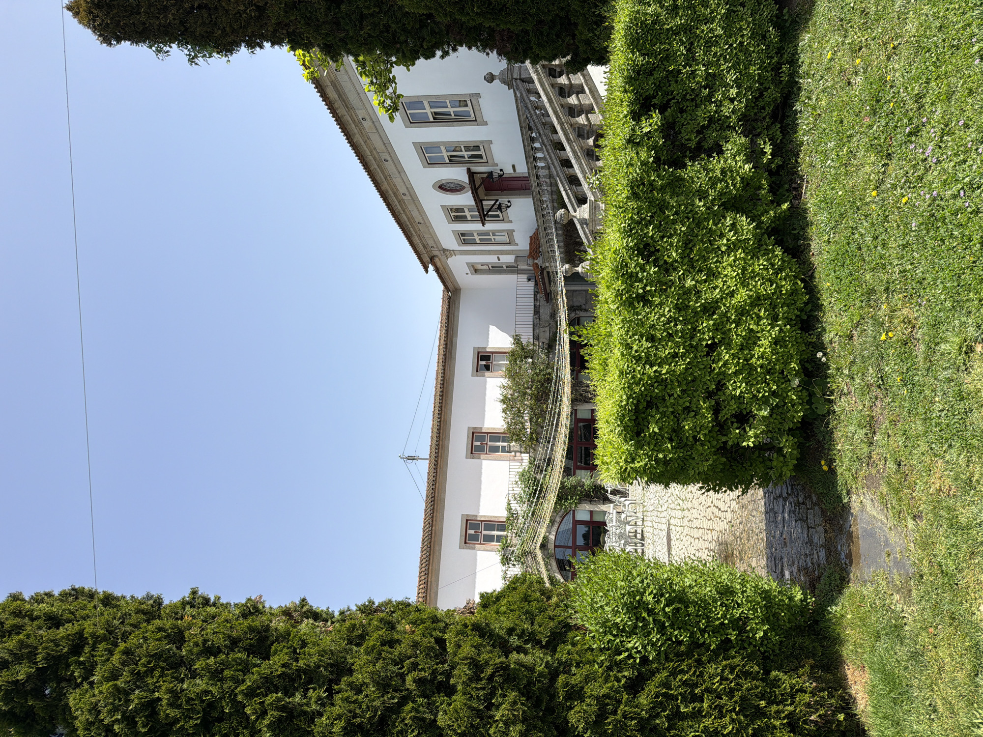 The image shows a building and garden in Vila Real, Portugal. The building is white with multiple windows and a red door. A stone staircase with decorative balusters leads up the side of the building. The building is surrounded by green hedges and grass. A stone pathway runs through the garden. Overhead electrical lines are visible against the blue sky. Outdoor furniture such as tables and chairs are on the ground floor. The photo's perspective is from a slightly low angle, looking up at the building, and rotated counter-clockwise.