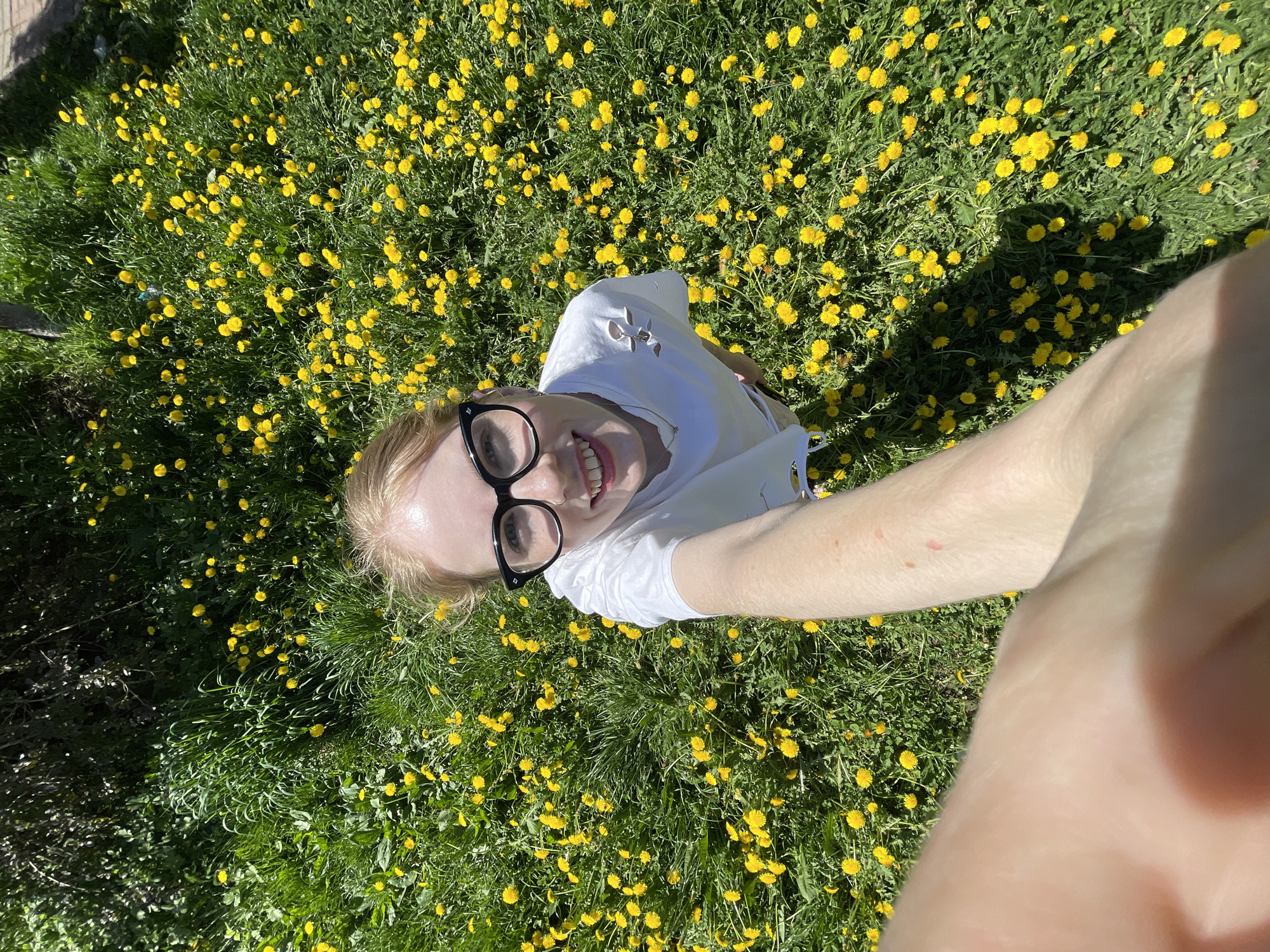 A young woman with fair skin, blond hair, and black-framed glasses lies on her back in a field of yellow flowers and green grass, possibly dandelions. She wears a white t-shirt and smiles at the camera, which she holds at arm's length, resulting in a selfie-style photograph taken from a low angle. The flowers and grass fill most of the frame, providing a vibrant background. A small portion of a dark-colored fence is visible in the top left corner. The location of the photograph is unknown and not necessarily Izhevsk, Russia.
