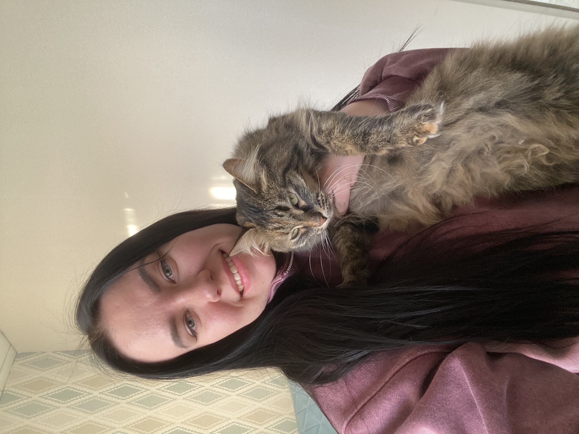A woman with long black hair and fair skin is holding a long-haired tabby cat. The woman is smiling and facing the camera. The cat is lying across the woman's upper chest and shoulder, with one paw resting on her upper arm. The woman is wearing a maroon-colored hooded sweatshirt. The background shows a beige ceiling and a wall with a light-colored geometric patterned wallpaper. Based on visual clues only, there is not enough information to determine that the image was taken in Chaikovsky, Russia.