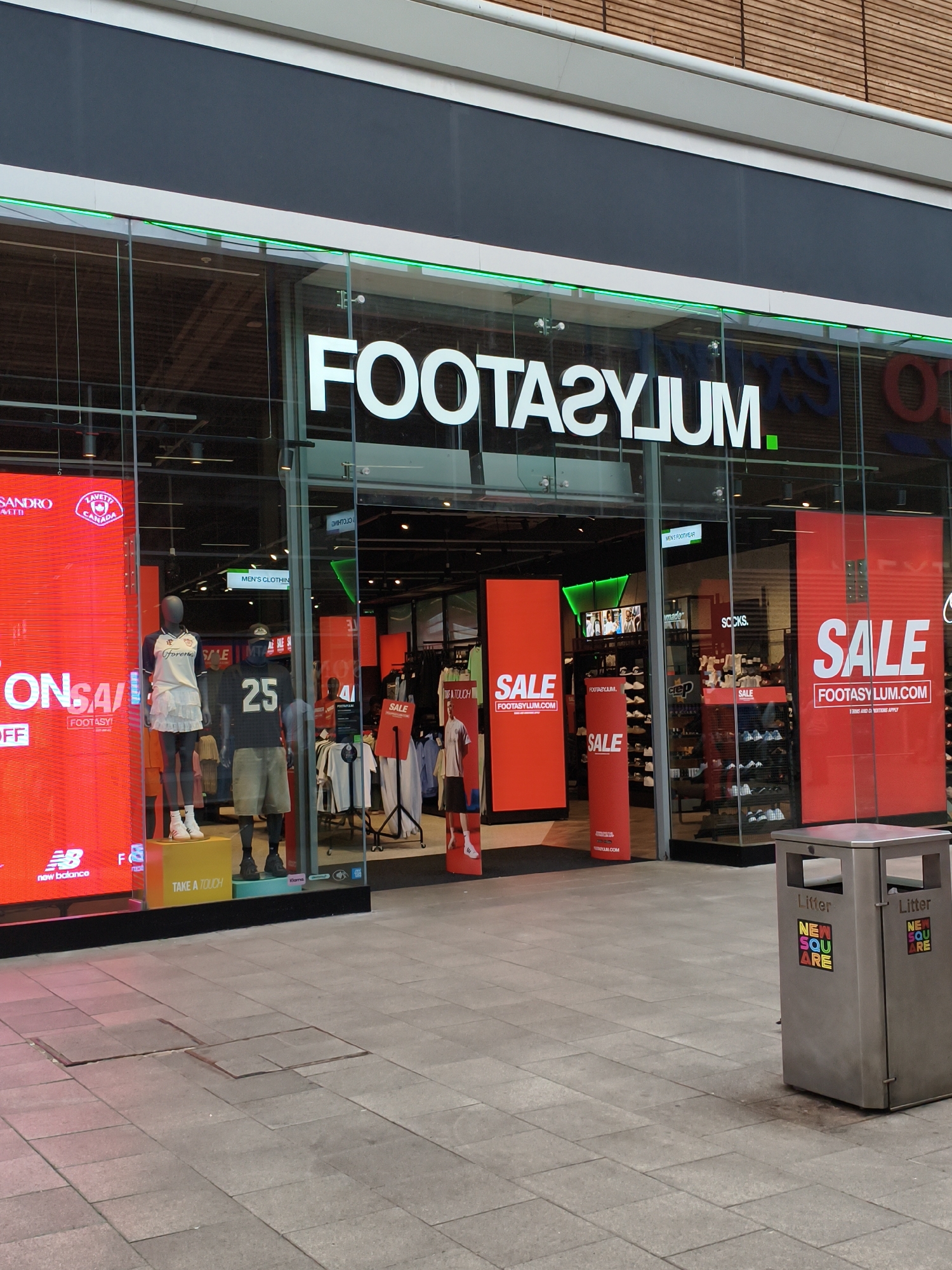 This is a medium, eye-level, exterior shot of a Footasylum store, located in Sandwell, United Kingdom. The store has a large glass storefront with the store name, "FOOTASYLUM," prominently displayed in large white lettering across the top facade. Green LED lighting runs along the top of the storefront. Inside the store, red banners with "SALE" are visible, advertising discounts. Two mannequins are displayed in the window, one wearing a white football jersey and white skirt, and the other wearing a black football jersey with the number "25" and khaki shorts. Various sports apparel and footwear are displayed on shelves and racks inside the store. To the right of the store entrance, a silver double-bin litter bin is situated on the paved ground. The bin has two openings and a sticker on each side with the words "NEW SQUARE" in colorful letters. The ground outside the store is paved with square tiles. The overall lighting suggests it is daytime, with some overcast conditions.