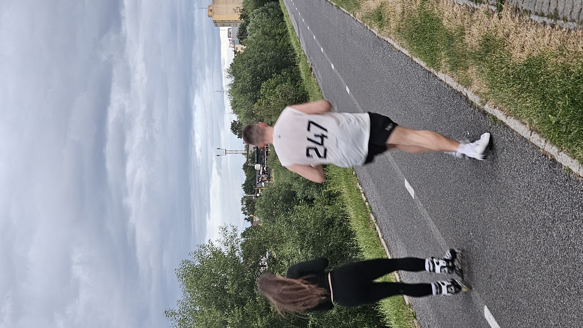 The image shows a paved pathway bordered by grassy verges and lush green trees. In the background, buildings and communication towers are visible, hinting at an urban or semi-urban setting, possibly within Prešov, Slovakia, given the context. On the pathway, two individuals are engaged in physical activity. On the left, a person with long, dark hair is rollerblading, wearing black leggings and a black cropped top. Their rollerblades are black with white accents. On the right, a person is running, seen from the back. They are wearing a white sleeveless t-shirt with the number "247" printed in black on the back, black shorts, white socks, and white running shoes. The sky is overcast with heavy grey clouds. The lighting suggests it is daytime, possibly late afternoon or early evening. The overall scene depicts a recreational activity taking place on an outdoor path.