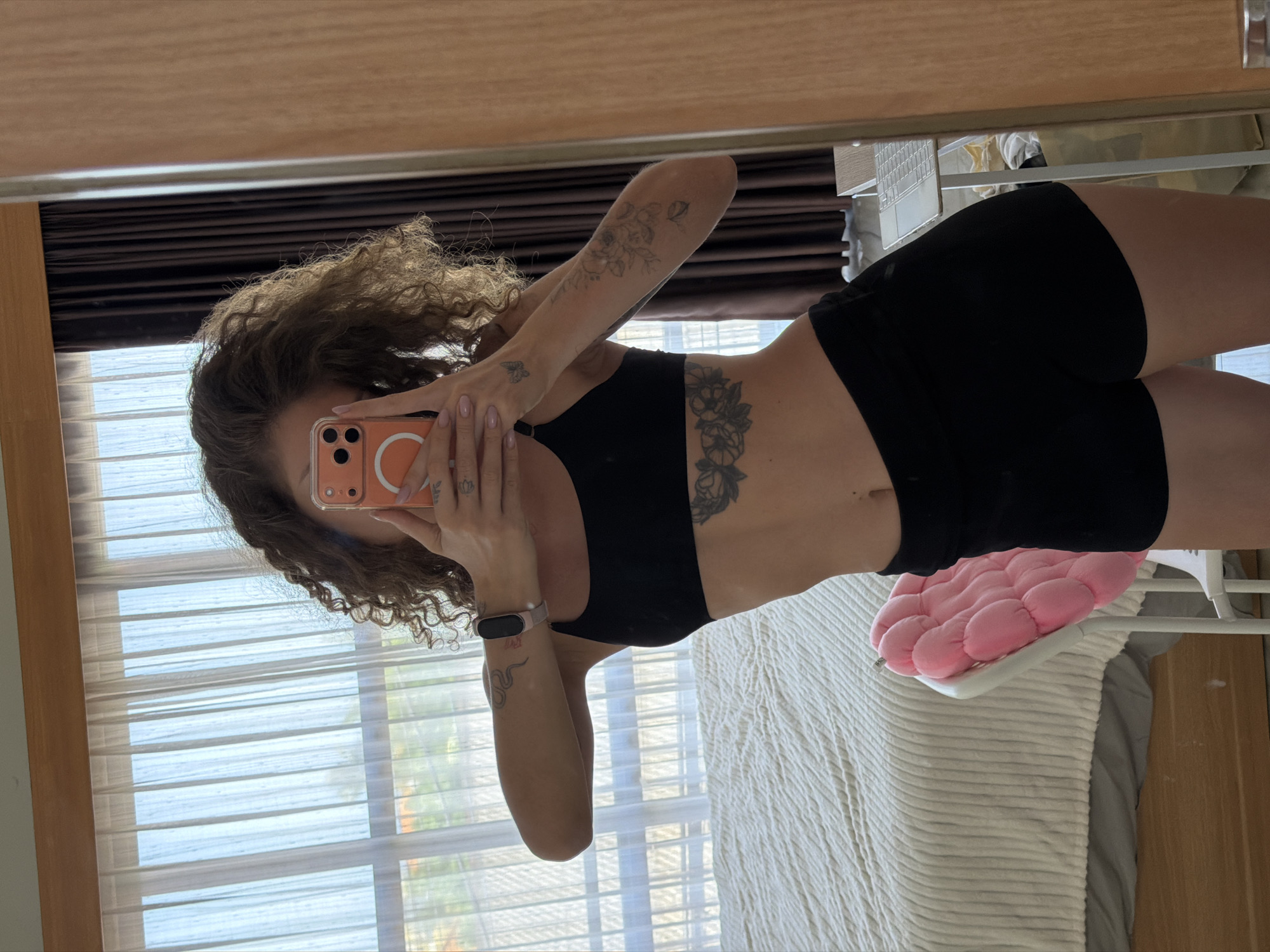 In a bedroom in North Kuta, Indonesia, a woman with curly brown hair takes a mirror selfie. She is wearing a black sports bra and black shorts. Her left arm, which is bent at the elbow, is holding an orange-cased smartphone to take the photo. She has tattoos on her left arm and a floral tattoo on her abdomen. A smartwatch is visible on her left wrist. In the background, sheer white curtains hang over a window, revealing a glimpse of the outdoors. A large bed with white bedding is to the left, and a pink plush cushion is on a white chair to the right of the bed. A wooden-framed mirror occupies the right side of the image, reflecting the scene.