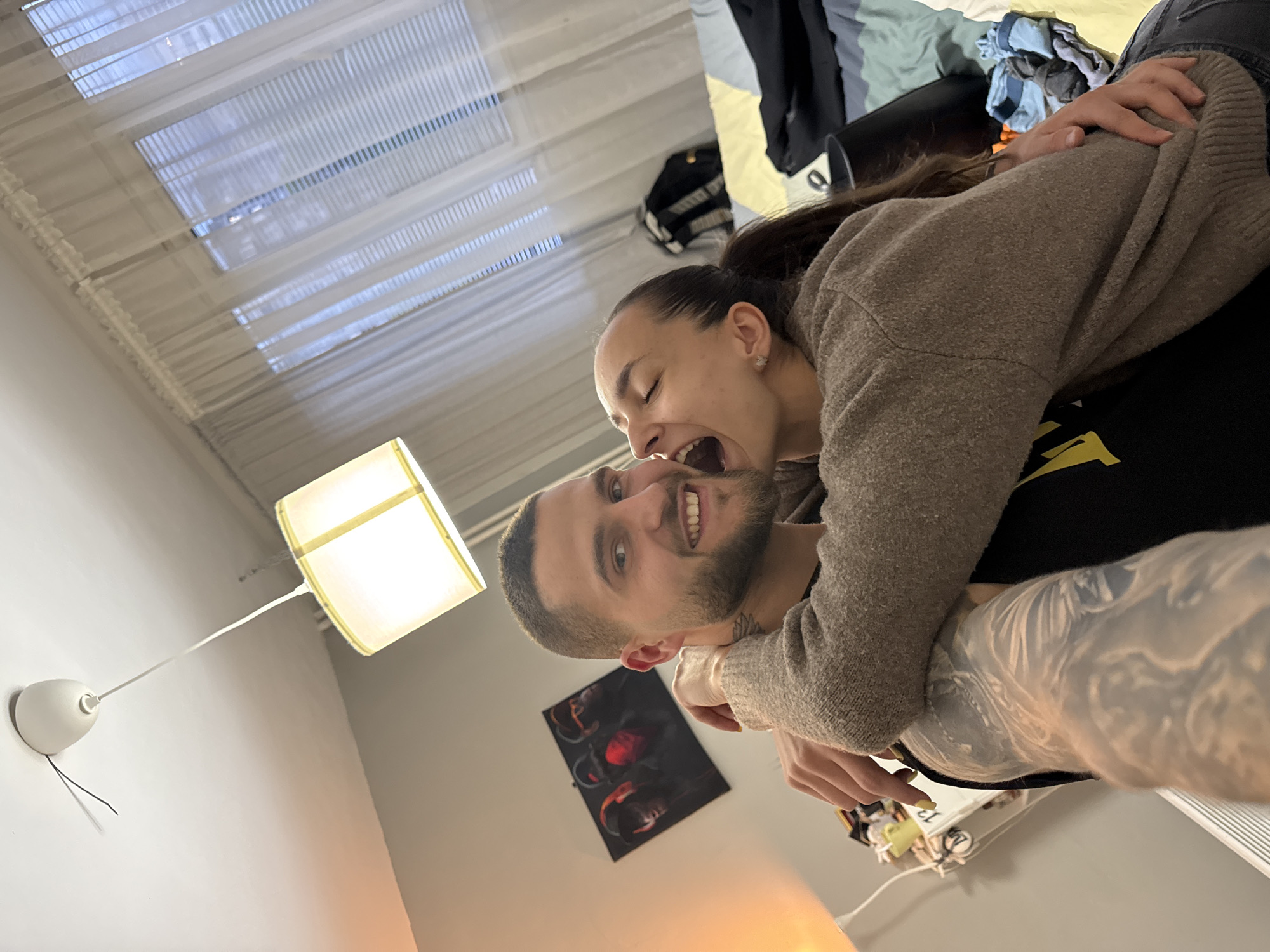 This indoor photograph captures a candid and joyful moment between a couple in what appears to be a cozy living space or bedroom in Sofia, Bulgaria. The image is taken from a low, dynamic angle.
In the foreground, a man with a short beard and hair smiles broadly at the camera. He is wearing a light brown knitted sweater, and a tattoo is visible on his neck. Embracing him from behind, a woman with dark hair tied back in a ponytail rests her head on his shoulder, her mouth open wide as if in mid-laugh or a playful "bite," her eyes closed in amusement. She also wears a similar light brown knitted sweater, and her arm, wrapped around the man, features an extensive tattoo sleeve. Both individuals convey strong emotions of happiness and playful affection.
The background includes a window covered by white horizontal blinds, allowing diffused light or privacy. A modern floor lamp with a square white fabric shade on the left emits a warm, inviting glow, suggesting either evening or reliance on artificial lighting. Below it, a smaller white wall-mounted light fixture with a visible black wire is present. In the upper right corner, a dark piece of furniture, possibly a bed or chair, is partially visible, with some clothes (including a light blue and white striped garment) casually placed on it. On the wall behind the man, a dark-framed picture featuring what appears to be red sneakers is hung. Various small items and wires are partially visible on a surface in the bottom right. No discernible text is visible in the image.