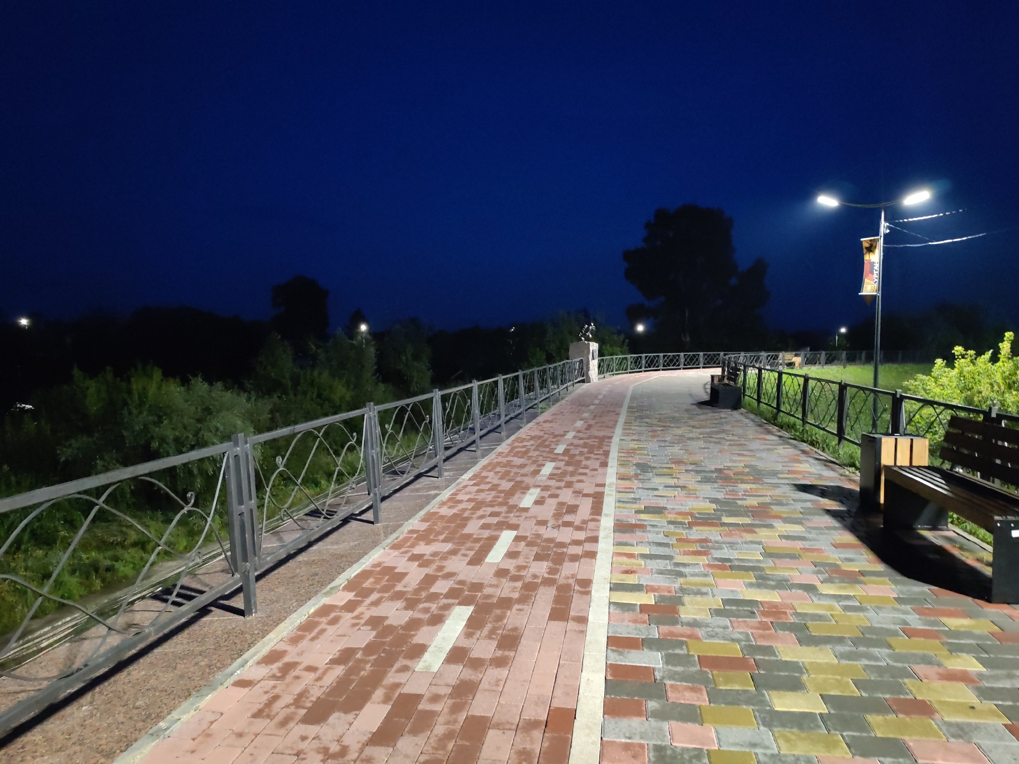 The image shows a paved walkway at night, illuminated by a streetlamp. The walkway is bordered by a metal fence on the left and a wooden bench on the right. The sky is dark blue, and the surrounding area is filled with trees and vegetation. A banner hangs from the lamppost. The walkway itself is made of colorful rectangular pavers, with a contrasting lighter colored line running down the center.