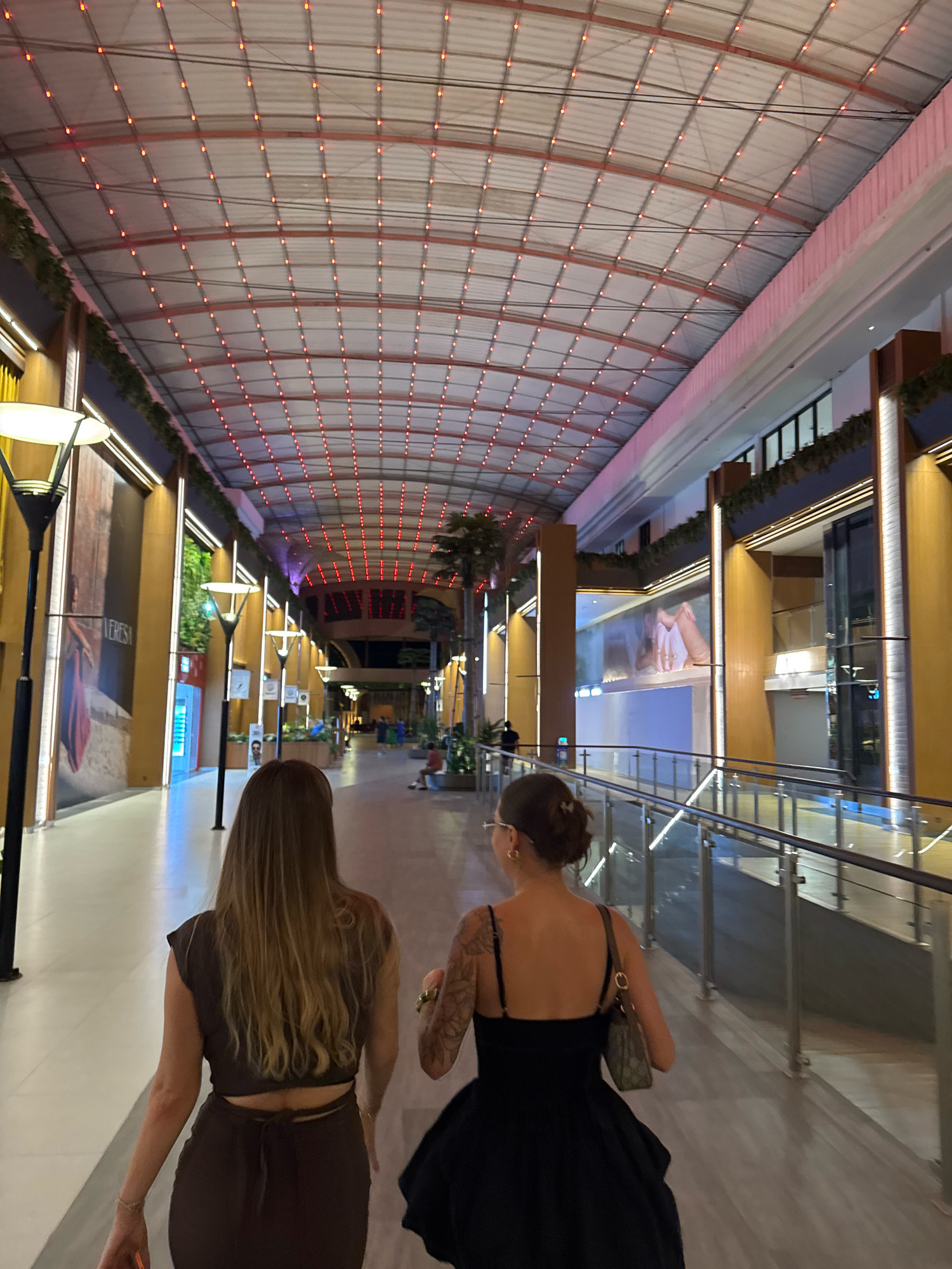 The image shows two women walking down a covered outdoor walkway in Kuta, Indonesia. The walkway has a curved, paneled ceiling with numerous small red lights strung across it. The sides of the walkway are lined with storefronts and illuminated signage. On the left, there are large posters and vertical lighting fixtures. On the right, a glass railing overlooks a lower level. Palm trees are visible in the background. The two women are walking away from the camera. The woman on the left has long blonde hair and is wearing a brown sleeveless top and skirt. The woman on the right has her hair in a bun and is wearing a black spaghetti-strap dress and carrying a handbag. Both appear to be in good spirits. The overall impression is a vibrant evening scene in a commercial area.