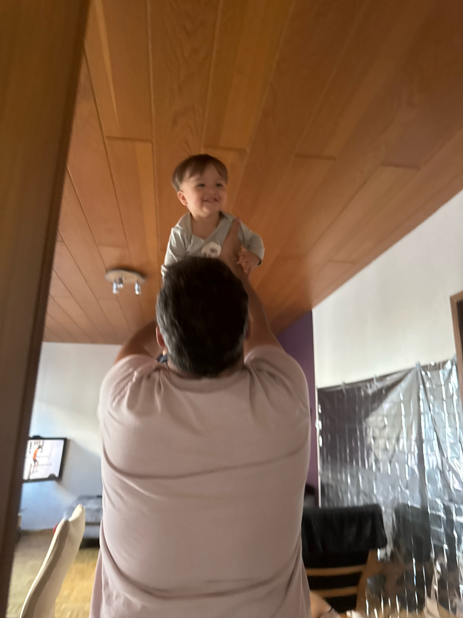 An adult, facing away from the camera, holds a smiling baby aloft in what appears to be an indoor residential setting. The adult wears a light pink t-shirt, and the baby is dressed in a light blue onesie. The background features a wooden slatted ceiling and walls, with a light fixture visible on the ceiling. To the right, a large silver metallic sheet is propped up against a wall, and a portion of a television screen displaying a person can be seen on the left. The perspective suggests the photo was taken from a low angle, looking upwards. The scene does not contain enough information to determine its location within Überherrn, Germany.