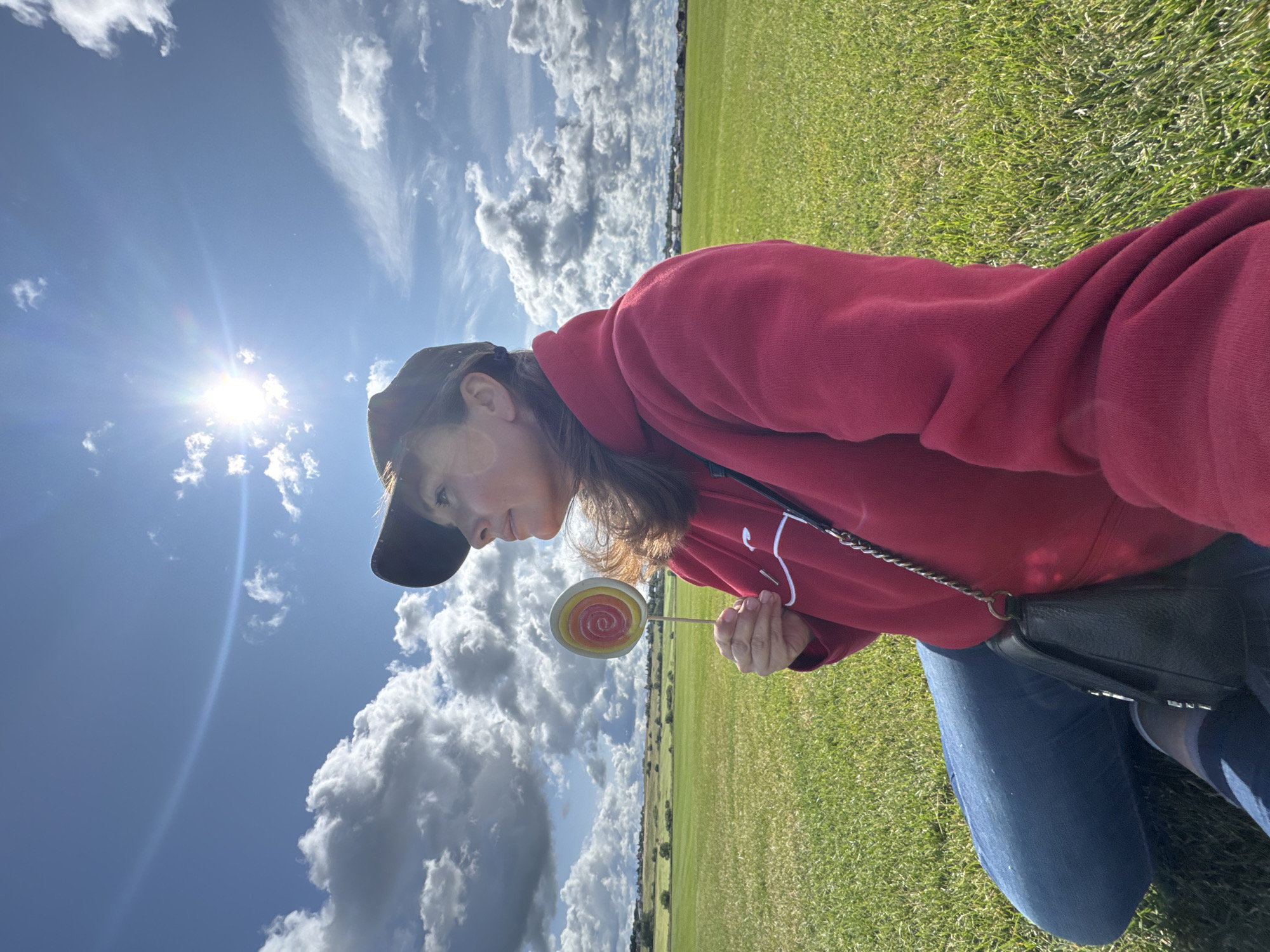 In Innolovo, Russia, a woman wearing a red hoodie and blue jeans sits in a grassy field under a bright, sunny sky with scattered clouds. She holds a lollipop with a spiral design of red, yellow, and white. She is also wearing a dark baseball cap and a black purse hangs from her shoulder. The background shows a gentle, green landscape with trees and some distant buildings.