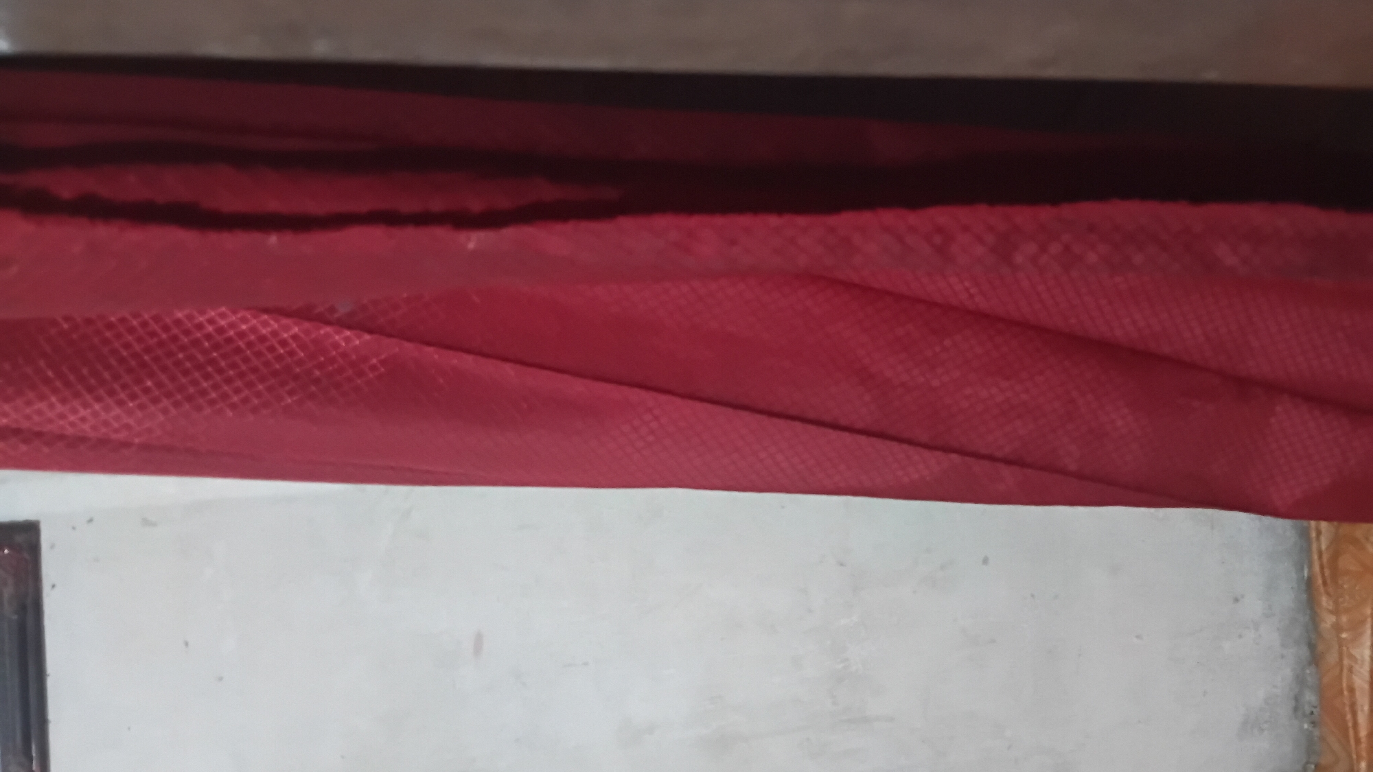 A full-frame, vertical, close-up shot shows a red textured curtain to the right, partially obscuring a plain white wall to the left. The curtain exhibits a diamond-shaped pattern and drapes in folds, with a shadow cast on its right side. The wall appears to be concrete or plaster and has some minor imperfections. At the bottom edge of the frame, a portion of a tiled floor with an orange-brown pattern is visible. The lighting is dim, suggesting an indoor setting. There are no people or specific identifiable landmarks of Prabumulih, Indonesia, in the image.