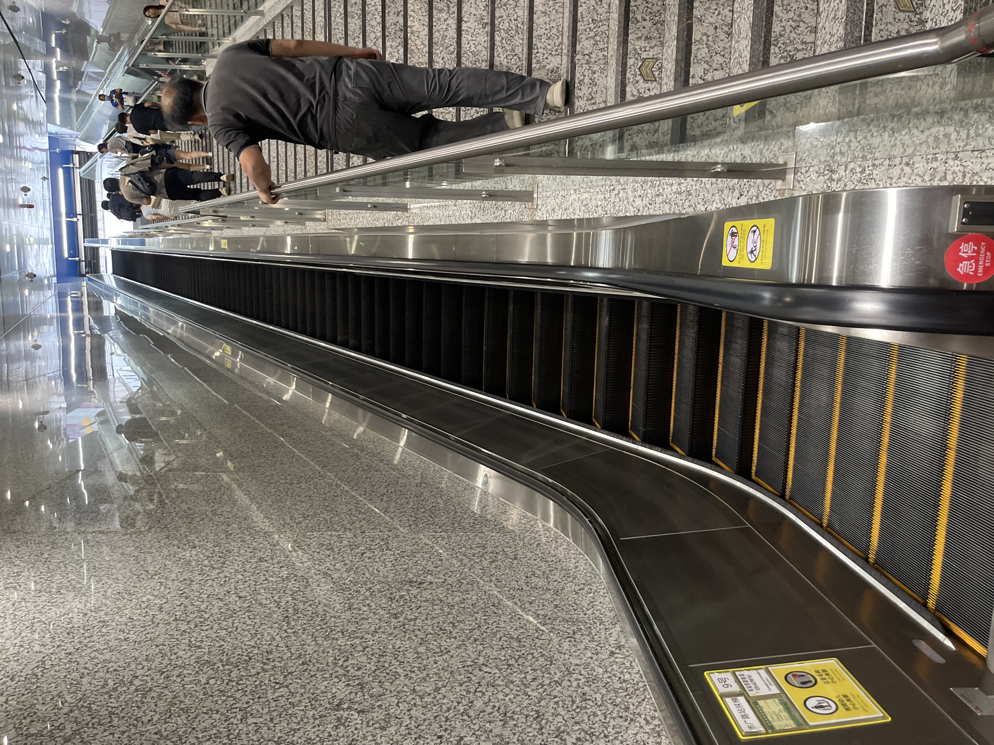 In a well-lit, modern subway station, an escalator moves downwards. Several people are present, with one man in the foreground walking down a set of stairs adjacent to the escalator. He is wearing a dark grey t-shirt and pants. Other individuals, some with backpacks, are also visible on the stairs and the escalator, suggesting a typical commute or travel scenario.
The scene is indoors, characterized by polished stone floors and metallic railings. The escalator itself is made of dark metal with yellow safety stripes on its steps. A large, reflective glass partition runs alongside the escalator and stairs, mirroring the environment.
Notable details include various warning signs. On the escalator, there are yellow signs with pictograms indicating "Careful of tripping" and "Beware of brushes," along with a red circular "EMERGENCY STOP" button. Another yellow sign near the bottom of the escalator displays Chinese characters and pictograms related to escalator safety. The overall impression is one of a functional and busy public transportation hub. The lighting suggests it is daytime, and there are no external weather cues. The location is identified by the text "市广路站扶梯 9号" which translates to "Shi Guang Road Station Escalator No. 9" in Panyu District, China. The presence of commuters indicates a normal operating day.