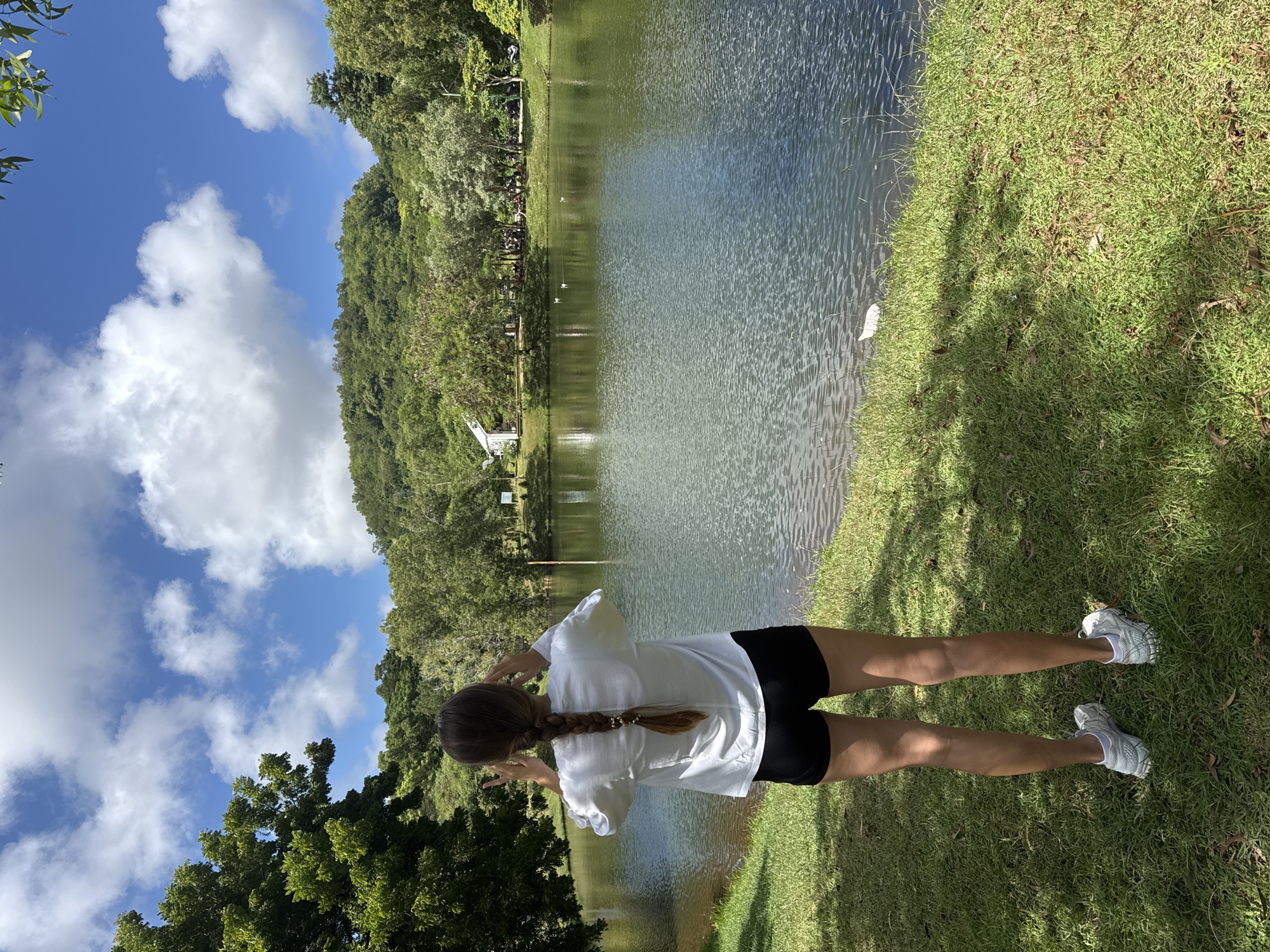The image shows a person standing on a grassy bank next to a lake on a sunny day with blue sky and scattered clouds. The person is wearing a white t-shirt and black shorts, and has their back to the camera. The lake is bordered by trees and foliage. On the opposite bank of the lake, there appears to be a small white building nestled among the trees. The exact location is Unknown, Unknown.
