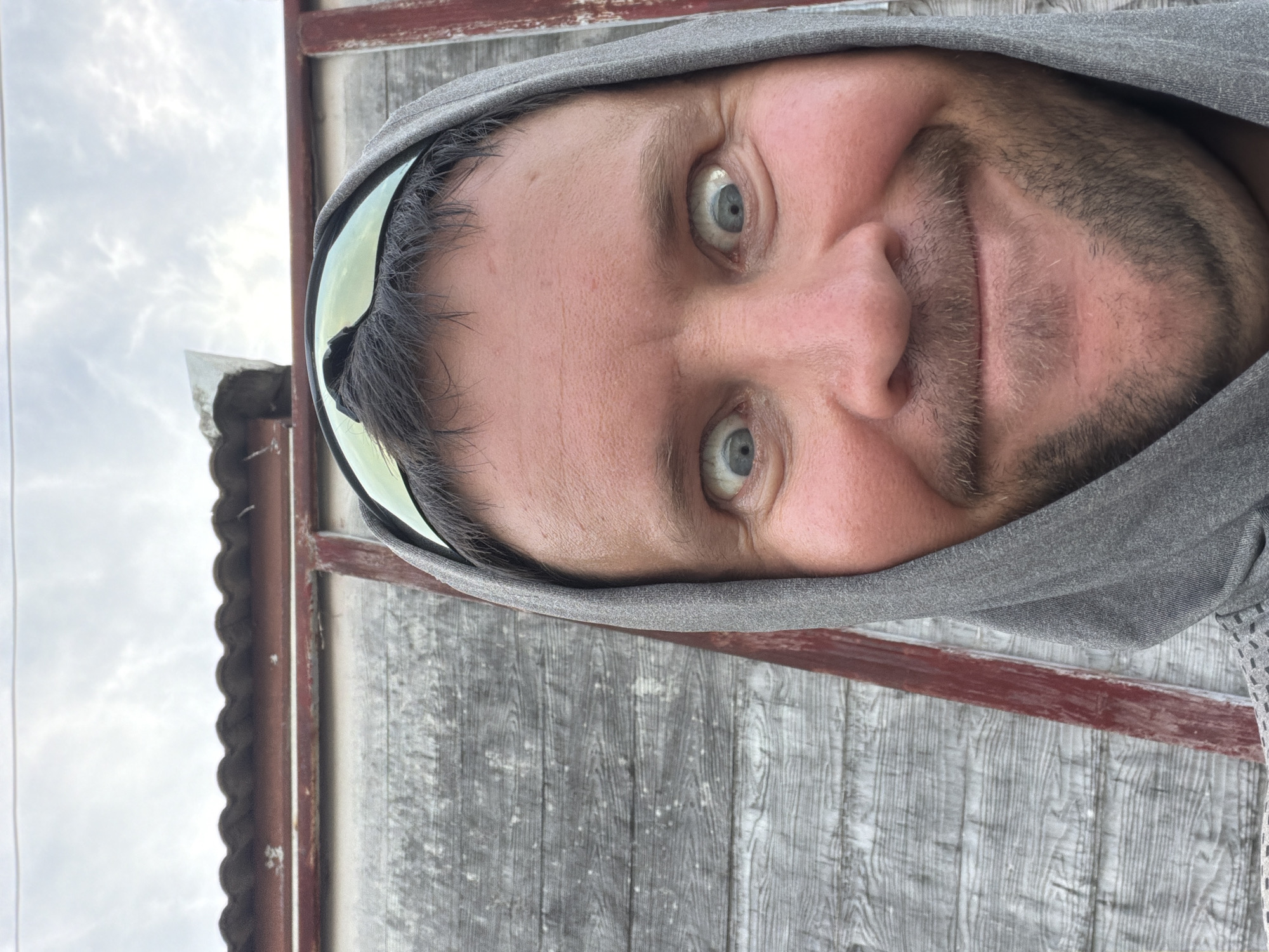 A man wearing a grey hooded sweatshirt and protective goggles is looking directly at the camera. His eyes are wide open and his mouth is in a wide smile. The background consists of a weathered wooden wall and a cloudy sky. The scene appears to be outdoors, possibly in Baan Mae Nam, Thailand, given the context.