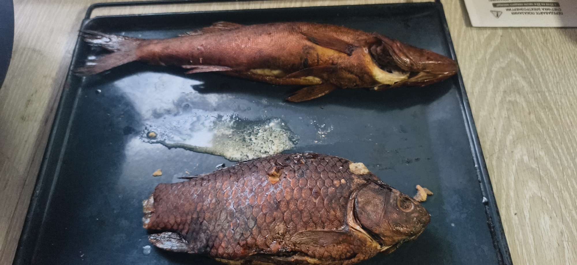 The image captures two cooked or smoked whole fish resting on a dark, rectangular metal baking tray. The fish are positioned diagonally across the tray. The upper fish is reddish-brown and elongated, with some lighter internal flesh visible. The lower fish is darker brown, broader, and shows distinct scales, possibly a carp. Around both fish, the tray has visible drippings, rendered fat, and solidified remnants from the cooking process.
The baking tray is placed on a light-colored wooden surface, suggesting an indoor setting like a kitchen or dining area. In the upper right corner, a portion of a white paper document is visible. It contains Cyrillic text, which includes "ПЕРЕДАВАЙТЕ ПОКАЗАНИЯ" (Submit readings), "СЧЕТЧИКА ЭЛЕКТРОЭНЕРГИИ" (Electricity meter), and "c 23-е до 25-е число каждого месяца" (from the 23rd to the 25th of each month). This text indicates a utility-related notice or bill.
No people are visible, and no specific activity beyond the presentation of the cooked fish is depicted. The lighting suggests an indoor setting during daylight hours. The image focuses on the prepared food, likely in a domestic context in Фоки, Russia.