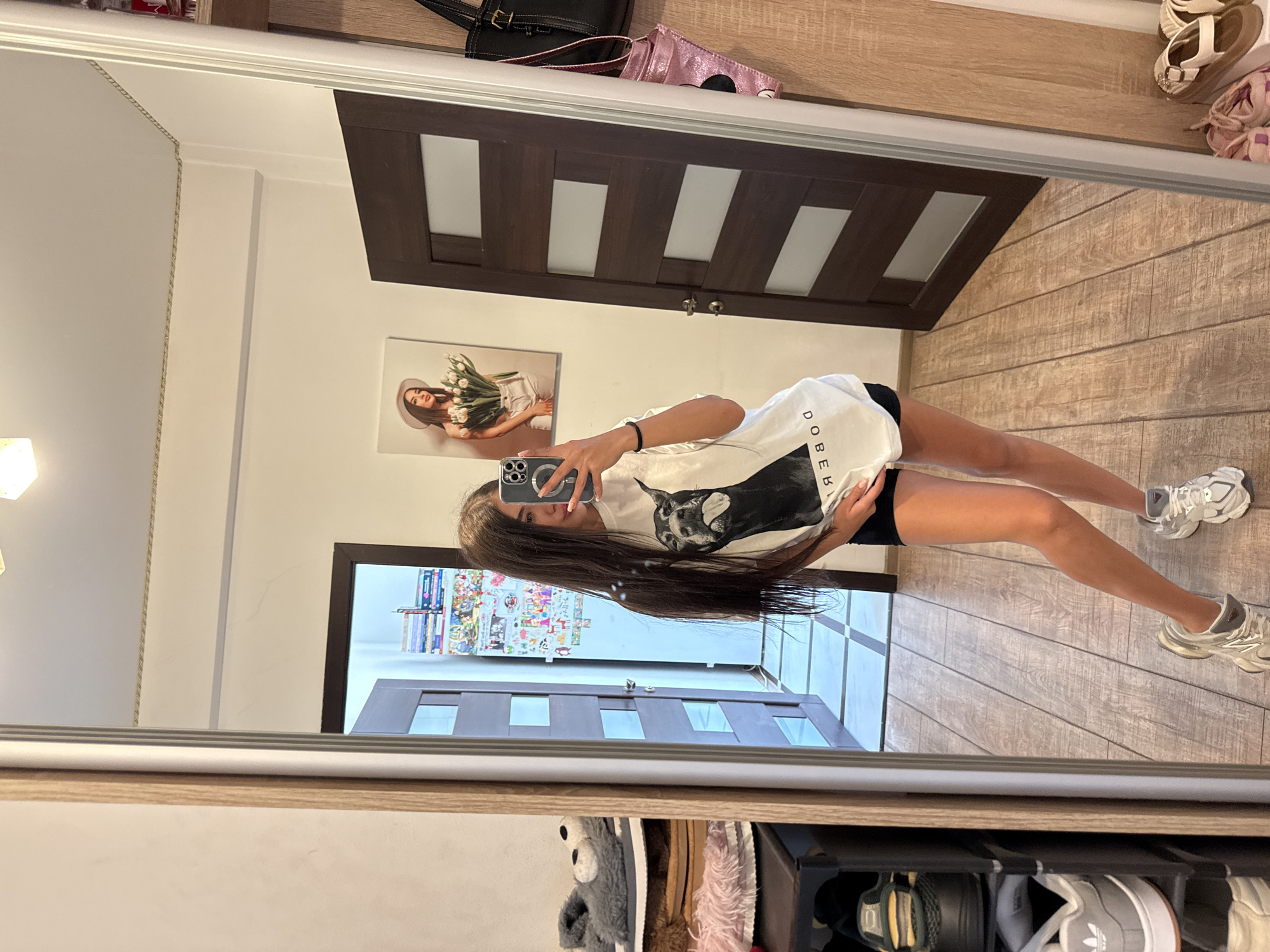 In a brightly lit room, a young woman with long, dark hair stands in front of a full-length mirror, taking a selfie with her phone. She is wearing a white t-shirt with a graphic of a dog and black shorts. Her hair is down, reaching past her shoulders. In the reflection of the mirror, several items are visible: a shoe rack filled with various footwear, a closed wooden door with frosted glass panels, and on the wall above, a framed picture of a woman holding tulips. A shelf unit to the right of the mirror displays bags and other accessories. The floor is made of wood-look tiles, and the walls are painted white. The overall impression is of a domestic space, possibly a bedroom or dressing area.