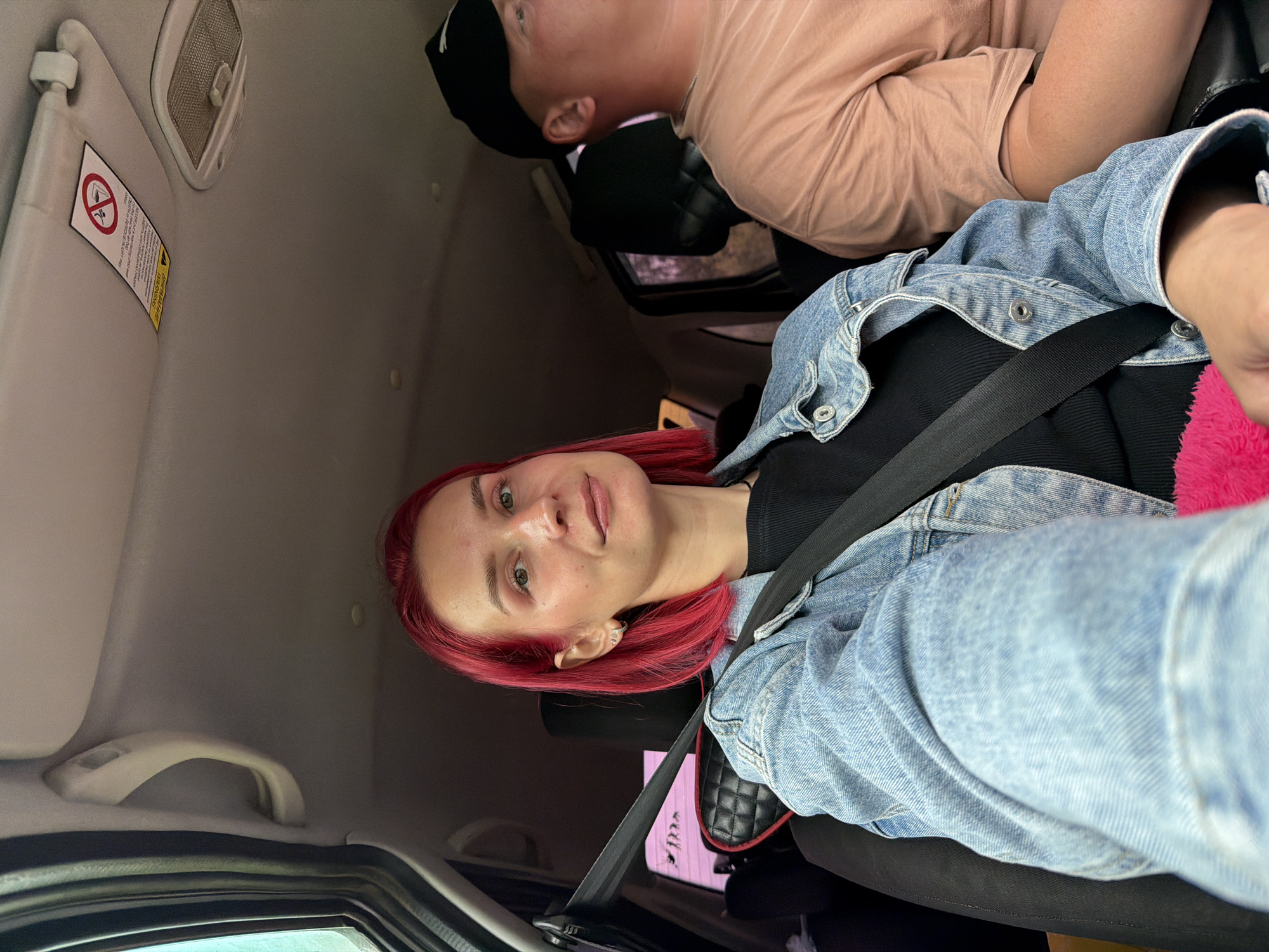 The image captures a selfie taken inside a car, featuring a woman in the foreground and a man in the background, set in what is contextually identified as Забегалово, Russia. The woman, with vibrant red hair, is in the front passenger seat, looking directly at the camera with a neutral expression. She wears a black top beneath a light blue denim jacket, with a black seatbelt fastened across her. A bright pink fuzzy object is partially visible to her right.
Behind her, a man is visible from the shoulders up, wearing a light peach-colored t-shirt and a black cap. He appears to be sleeping or resting against a car seat headrest. The car's interior is a light grey, showing the headliner, an overhead light, and a sun visor.
A prominent warning label on the sun visor features a red circular symbol depicting a child seat with an airbag, indicating "Do not place rear-facing child seat on front seat with active airbag." Below this symbol, the word "WARNING" is distinctly visible in yellow, accompanied by smaller text that includes phrases such as "SEVERE INJURY or DEATH can occur...". Other text on the label is present but not entirely legible.
The lighting suggests it is daytime. The overall scene conveys a casual moment, possibly during travel or a road trip.