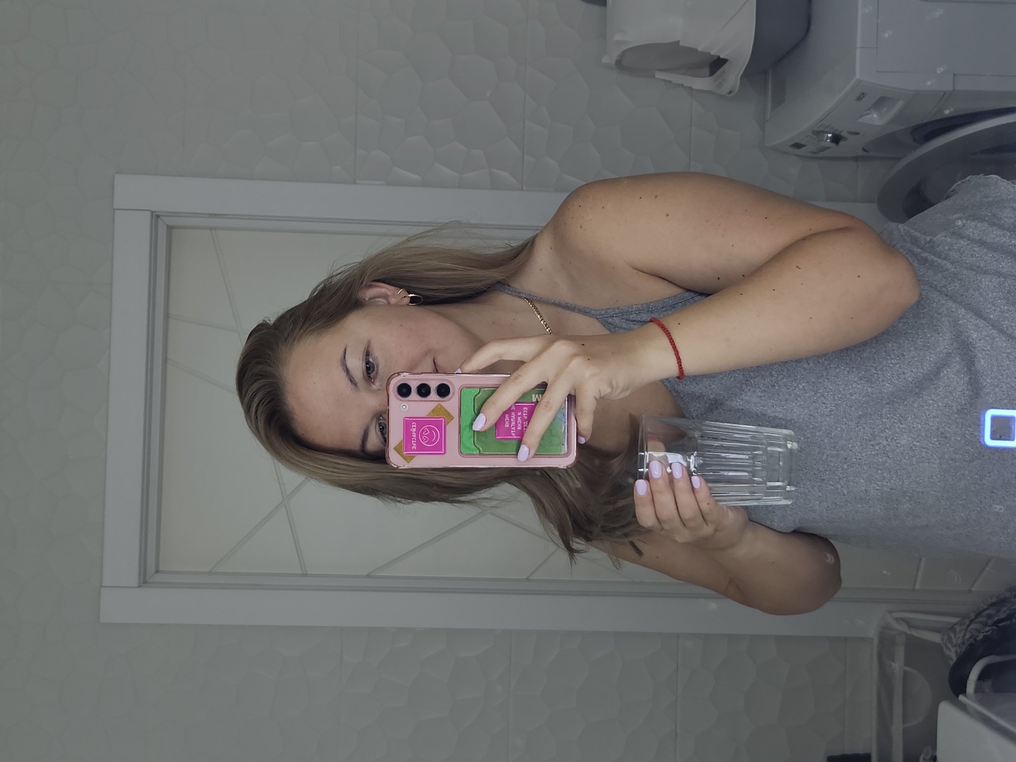 A medium shot captures a woman in a gray tank top taking a mirror selfie in a bathroom. She is holding a clear glass of water in her left hand and her right hand is holding up a pink smartphone with a transparent case and a pink cardholder attachment. Her blonde hair is partially pulled back. She is wearing small gold hoop earrings and a thin gold necklace. A thin red string bracelet is on her left wrist. The bathroom features white, textured tiles on the walls and a white framed door with geometric paneling. A washing machine and a cat litter box are visible in the background to the right. The image was taken in a bathroom in Rostov-on-Don, Russia.