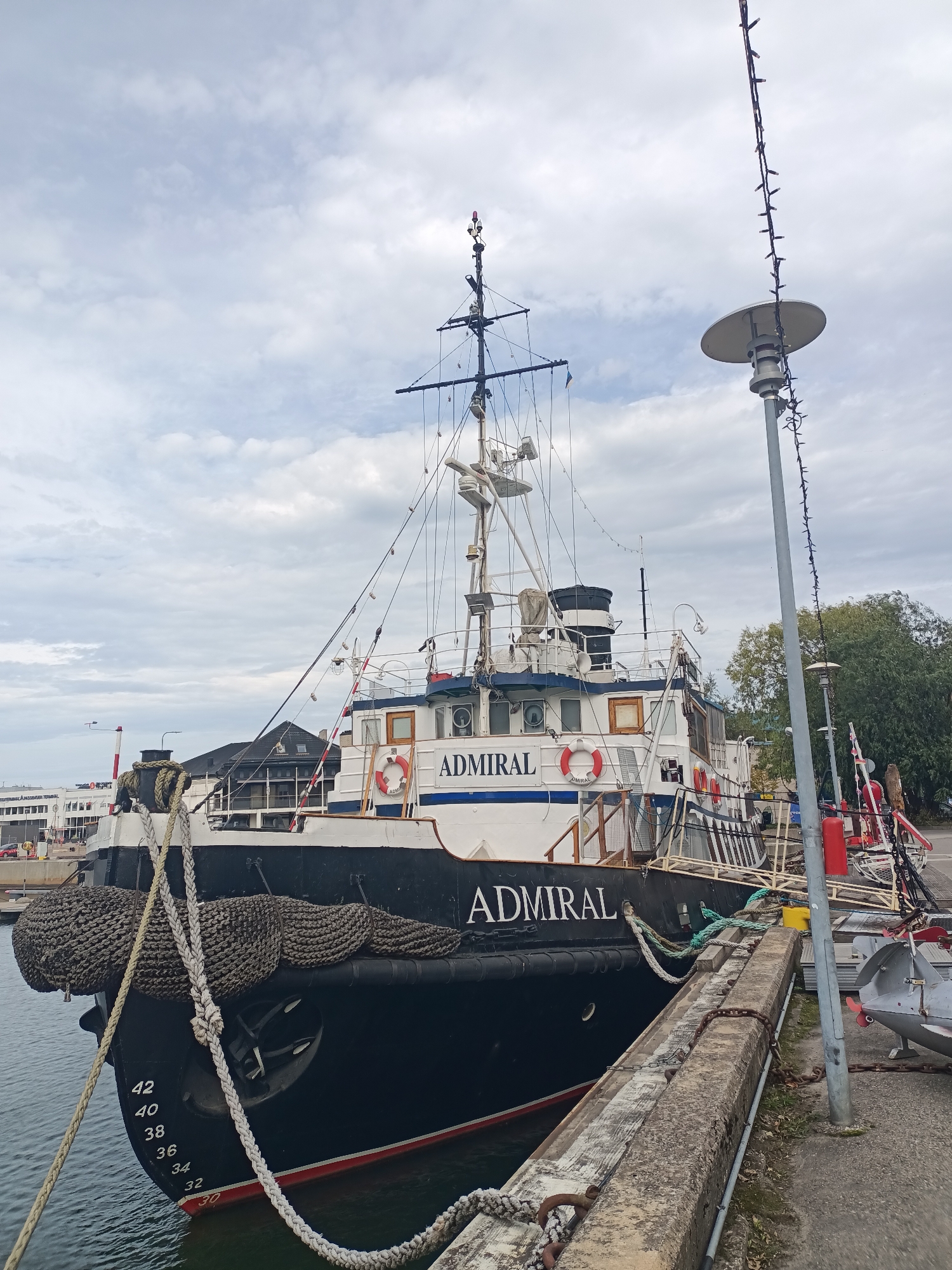 The image shows a ship named "ADMIRAL" docked at a harbor in Tallinn, Estonia. The ship is black and white with the name "ADMIRAL" clearly visible on its side in large white letters. Several life preservers, also with "ADMIRAL" written on them, are attached to the ship's railing. The ship is secured to a concrete dock with thick ropes. On the side of the ship, depth markers are visible, with numbers ranging from 30 to 42. In the background, there is a cloudy sky, some trees, a lamppost adorned with lights, and buildings that appear to be part of a port or terminal. The overall scene depicts a maritime setting in a city.