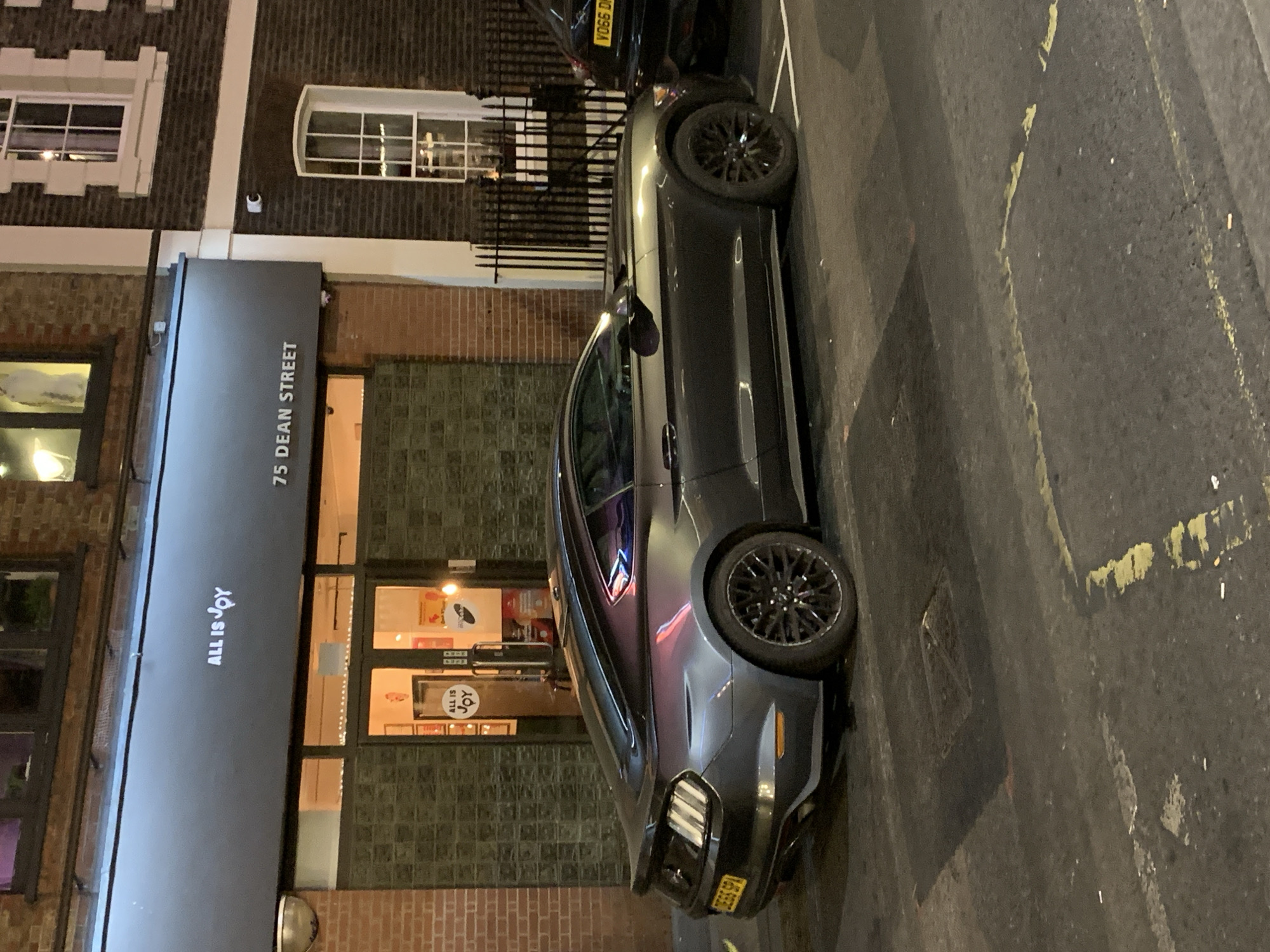 The image shows a gray Ford Mustang parked on a street in front of a building. The building's awning reads "75 Dean Street" and "All is Joy". Dean Street is located in the city of London, United Kingdom. The car is parked on the right side of the street, facing away from the viewer. There is another car parked partially visible ahead of the Mustang. The street appears to be wet. The photograph was likely taken at night.