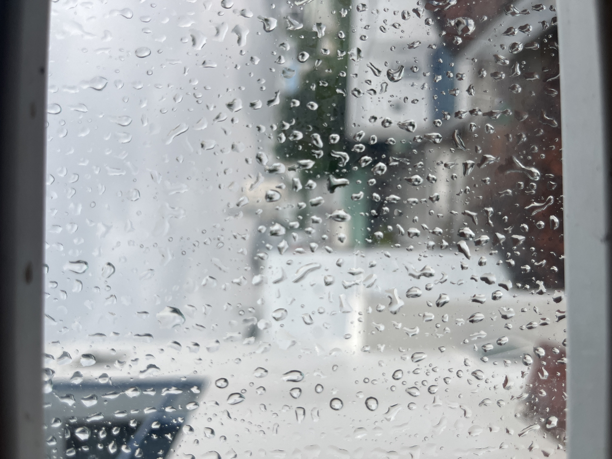 View from a window covered in raindrops. The blurred background shows a street scene, with hints of buildings and greenery, possibly located in Bac Nha Trang Ward, Vietnam.