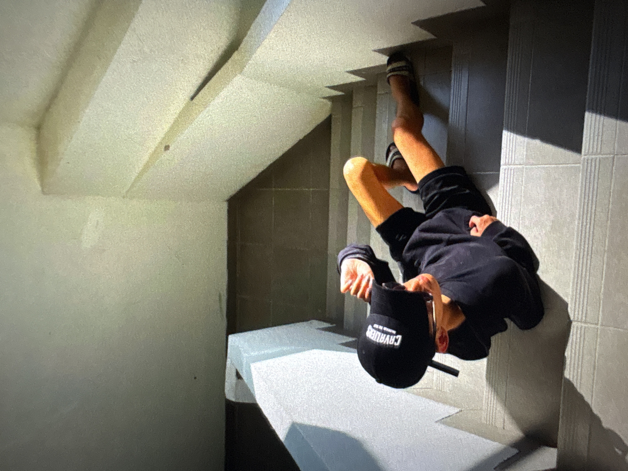 An overhead shot shows a person wearing a black baseball cap and black shorts, lying on their side on a set of concrete stairs. The person is wearing a black sweatshirt, and slides on their feet. The stairs are outdoors and appear to be part of a building with concrete walls. Sunlight casts shadows across the stairs. The image is taken in North Jakarta, Indonesia.